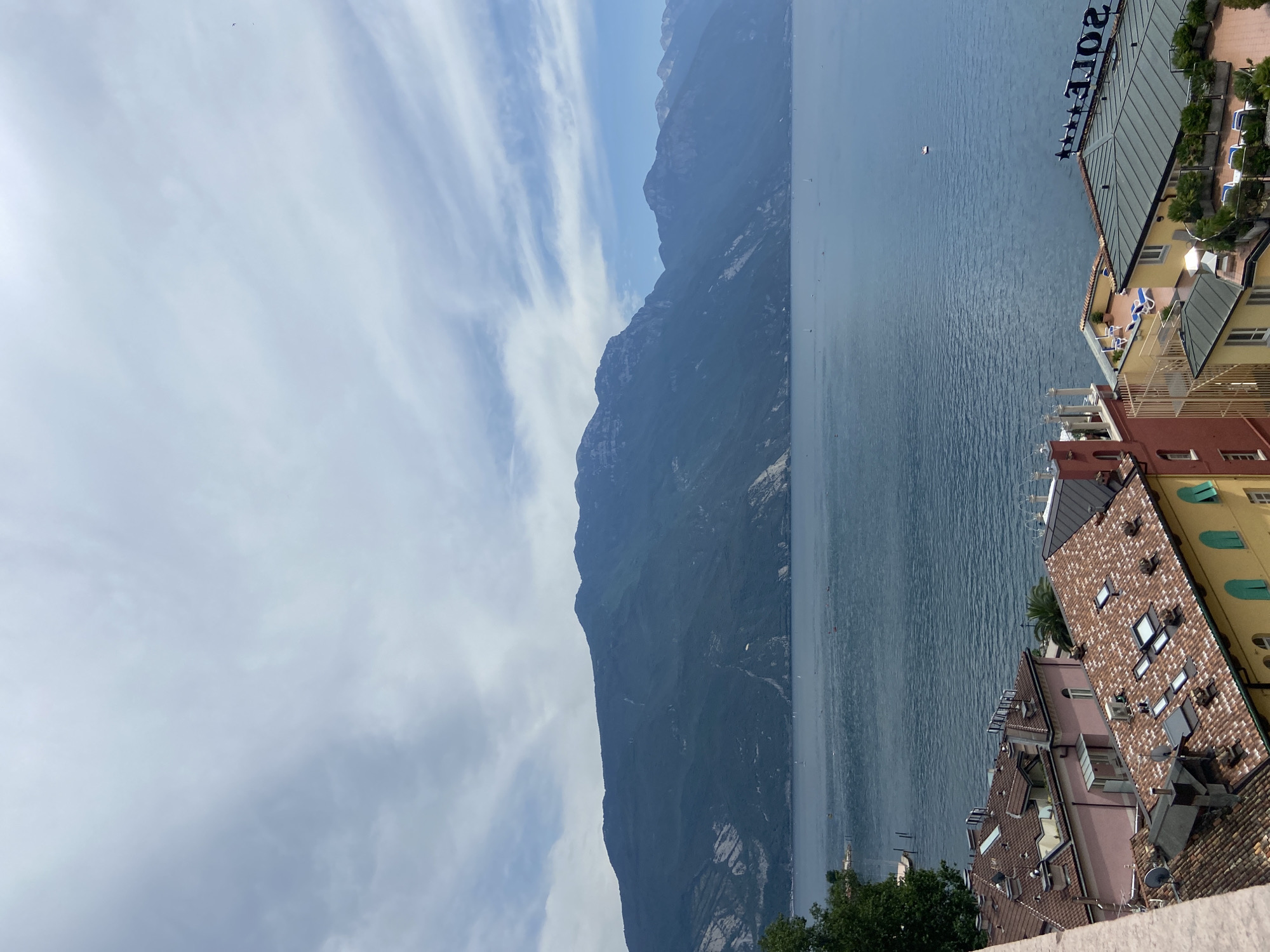 This is a high-angle, full shot of a vast blue lake with a mountainous backdrop, taken from a residential area. In the foreground, red-tiled roofs of buildings are visible, some with skylights, chimneys, and satellite dishes. One building has a pink facade, and another is bright yellow with green shutters. Rooftop patios with lounge chairs and plants can be seen. In the midground, the expansive blue lake is dotted with several small boats and a single white boat in the distance. The background is dominated by a large, dark, tree-covered mountain range that stretches across the horizon. The sky above is partially cloudy with streaks of white clouds against a pale blue sky. A paraglider is visible in the distance above the mountain. On a building in the far right, a sign reads "SOLE" in large letters. The scene suggests a clear, summery day in Riva del Garda, Italy, a popular tourist destination known for its lake and mountains.