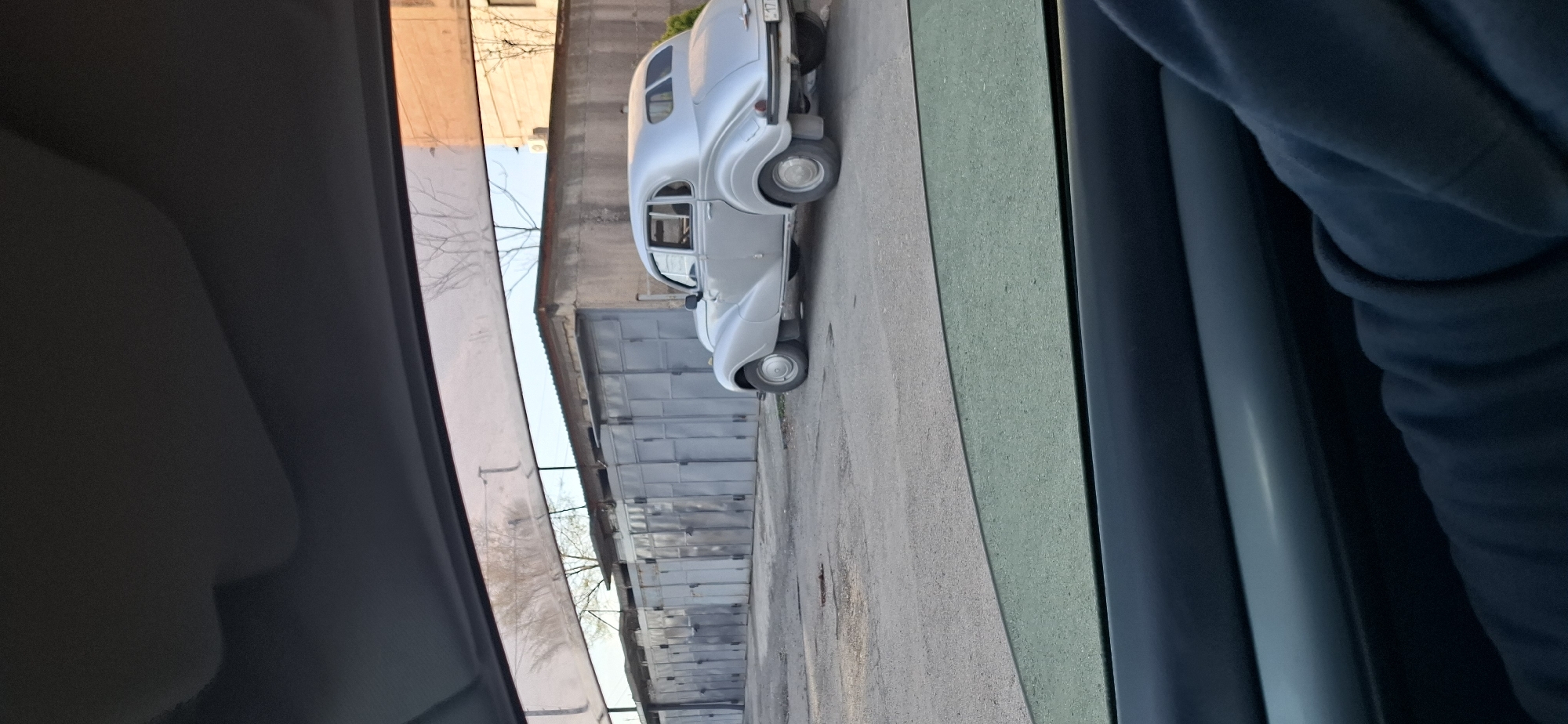 The image shows a light gray retro car parked near a row of gray garages. The photo is taken from inside another car, likely a vehicle's interior. The location appears to be an urban area in Yerevan, Armenia, based on the architectural style of the garages and general surroundings. There is a person in a blue hooded sweater partially visible within the capturing vehicle. The time of day seems to be daytime.