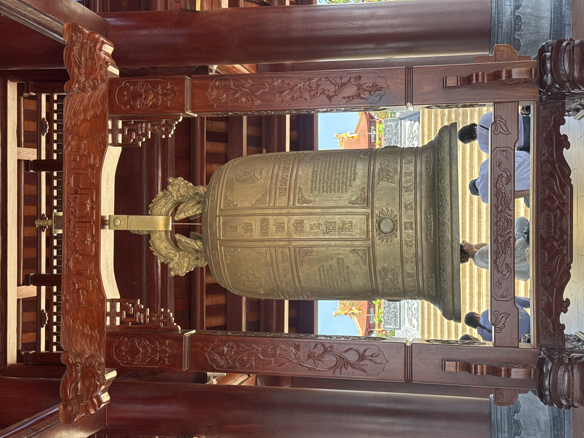 The image captures an interior view of a Vietnamese temple, likely in Diên Khánh, Vietnam. Dominating the scene is a large, ornate bronze bell, intricately decorated with carvings. The bell is suspended within a wooden structure, featuring detailed carvings of floral motifs and mythical creatures, suggesting traditional craftsmanship.
Several individuals are visible in the background, some appearing to be praying or participating in a religious ceremony. They are dressed in white, a common color for religious attire, and their posture suggests reverence. The bell itself has text inscribed on its surface in what appears to be Vietnamese script, though it is difficult to read clearly in the image. The lighting suggests it is daytime, with sunlight filtering through openings in the temple structure. The overall atmosphere is serene and spiritual. The wooden elements are a rich, dark brown, indicating aged or treated wood, and contrast with the metallic sheen of the bell. The architectural style points to a traditional Asian temple setting.