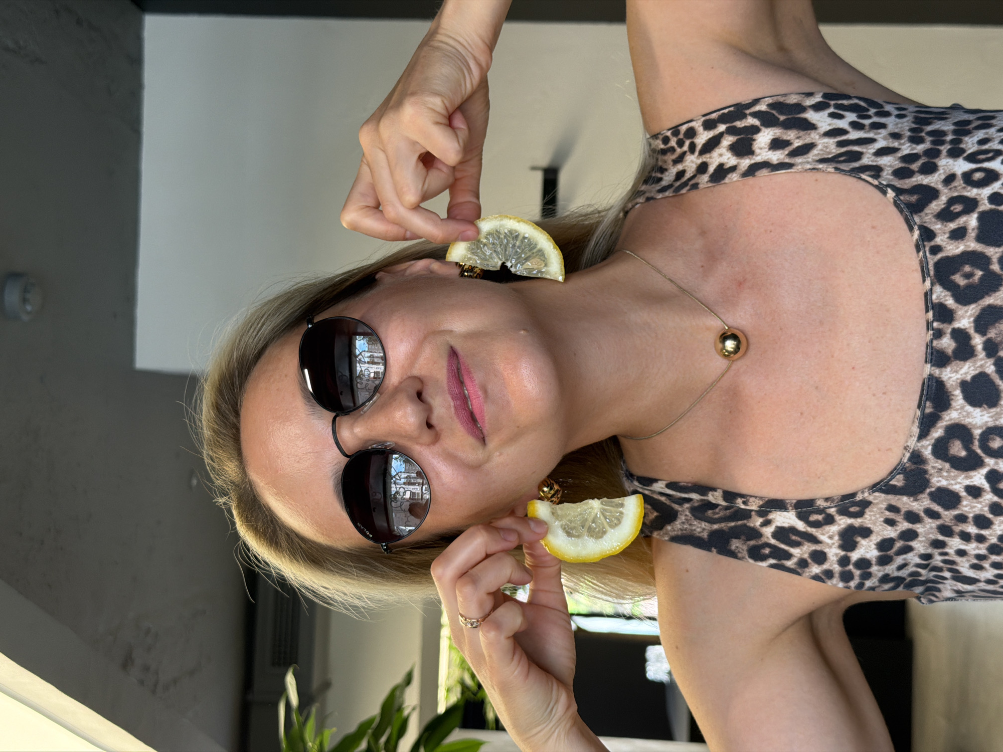 A woman with blonde hair and sunglasses is holding lemon slices as earrings. She is wearing a leopard print top and a gold necklace. The image appears to have been taken indoors, possibly in a cafe or restaurant, with blurred background elements. There is no information in the image to suggest a location in Izhevsk, Russia.