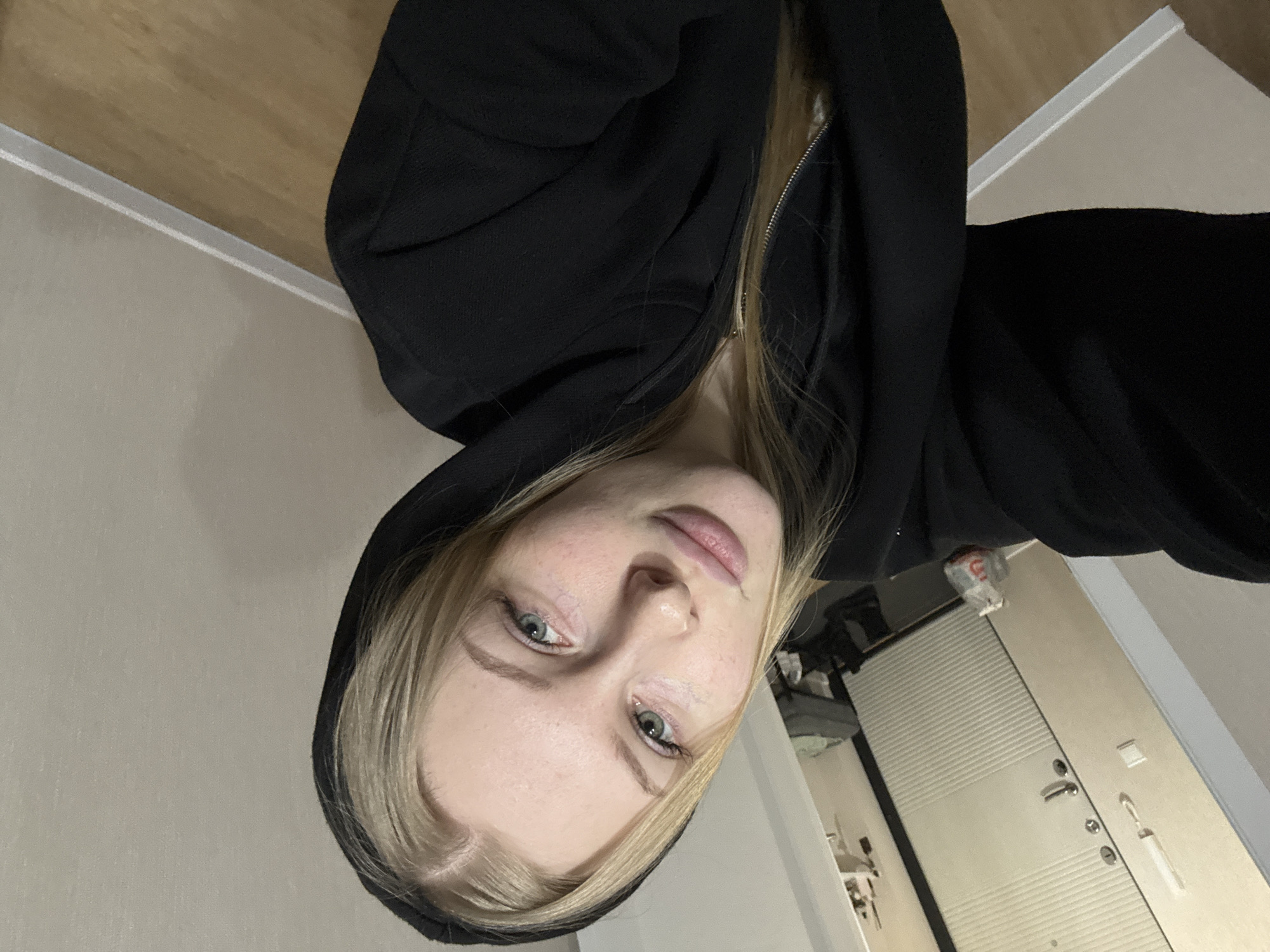 A close-up, portrait-oriented indoor selfie features a young woman with light blonde hair and blue-green eyes, looking directly at the camera. She has fair skin and visible freckles on her nose and cheeks. She is wearing a black hooded sweatshirt, partially zipped, with the hood pulled up around her head. Her expression is neutral and calm.
The background shows an interior space, likely a room or hallway. To her left (our right when viewing the image), a light-colored wall with a wooden floor or surface along the top edge is visible. To her right (our left), a light-colored door with horizontal panels, a silver handle, and multiple locks is prominent. A white light switch or power outlet and a small, white hanging item are on the wall next to the door. Above the door, a small red and white object is partially visible. The overall lighting suggests an indoor setting, possibly with artificial light. The architectural elements are consistent with a location such as Perm, Russia. No text is visible in the image.