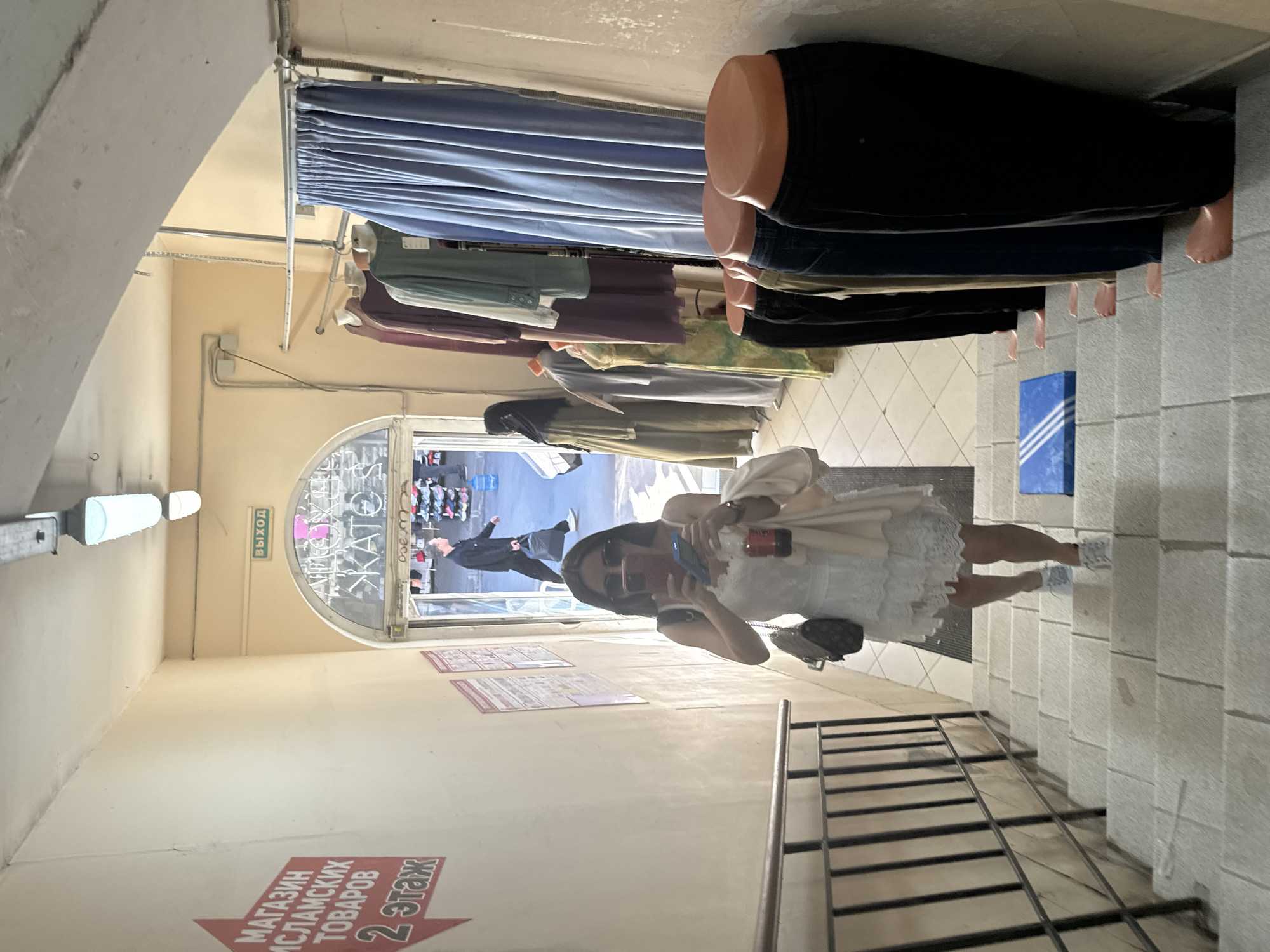 The image shows a woman taking a selfie in a hallway that appears to be part of a shopping area or mall in Saint Petersburg, Russia. The woman is wearing a white dress and sunglasses, and is holding a phone and a drink. She is standing on a tiled floor next to a staircase. In the background, there is a doorway leading to a shop with clothing racks and mannequins. There are signs visible in the hallway, one of which reads "МАГАЗИН ИСЛАМСКИХ ТОВАРОВ" (Islamic Goods Store) and indicates "2 ЭТАЖ" (2nd floor). Another sign above the doorway reads "ВЫХОД" (Exit). The lighting suggests it is daytime. The overall atmosphere appears casual and everyday.