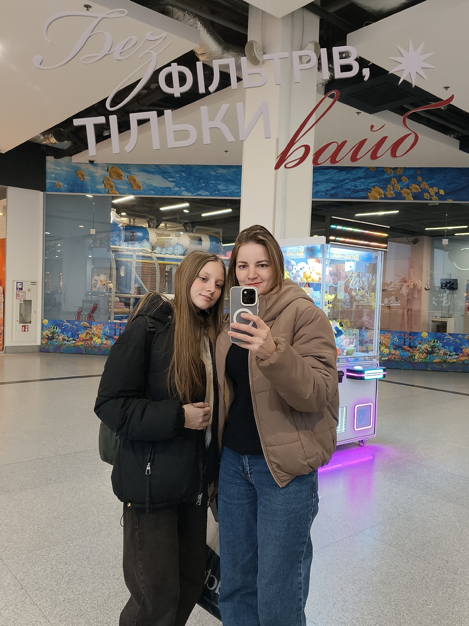 This full-shot image captures two females, an adult woman and a younger girl, taking a selfie in a brightly lit indoor environment, likely a shopping mall or entertainment center in Dnipro, Ukraine. The woman, positioned on the right, wears a light brown puffer jacket and blue jeans, holding a smartphone in her right hand to capture the reflection. The younger girl on the left wears a black puffer jacket and dark trousers, with long light brown hair. Both individuals look towards the camera with calm, subtle smiles.
The background reveals an entertainment area. To the left, behind the girl, is a children's play zone featuring large blue and white structures, adorned with illustrations of fish and an underwater theme. To the right, behind the woman, stands a white claw machine filled with plush toys, emitting colorful neon lights that cast a purple glow on the floor.
A prominent sign, reflected above the subjects, displays text in Ukrainian. The white letters read "Без ФІЛЬТРІВ, ТІЛЬКИ" (Bez FIL'TRIV, TIL'KY), meaning "Without FILTERS, ONLY". This is followed by a stylized red script word, "вайб" (vayb), which means "vibe." Together, the phrase translates to "No filters, just vibe." The setting suggests a casual outing or recreational activity.