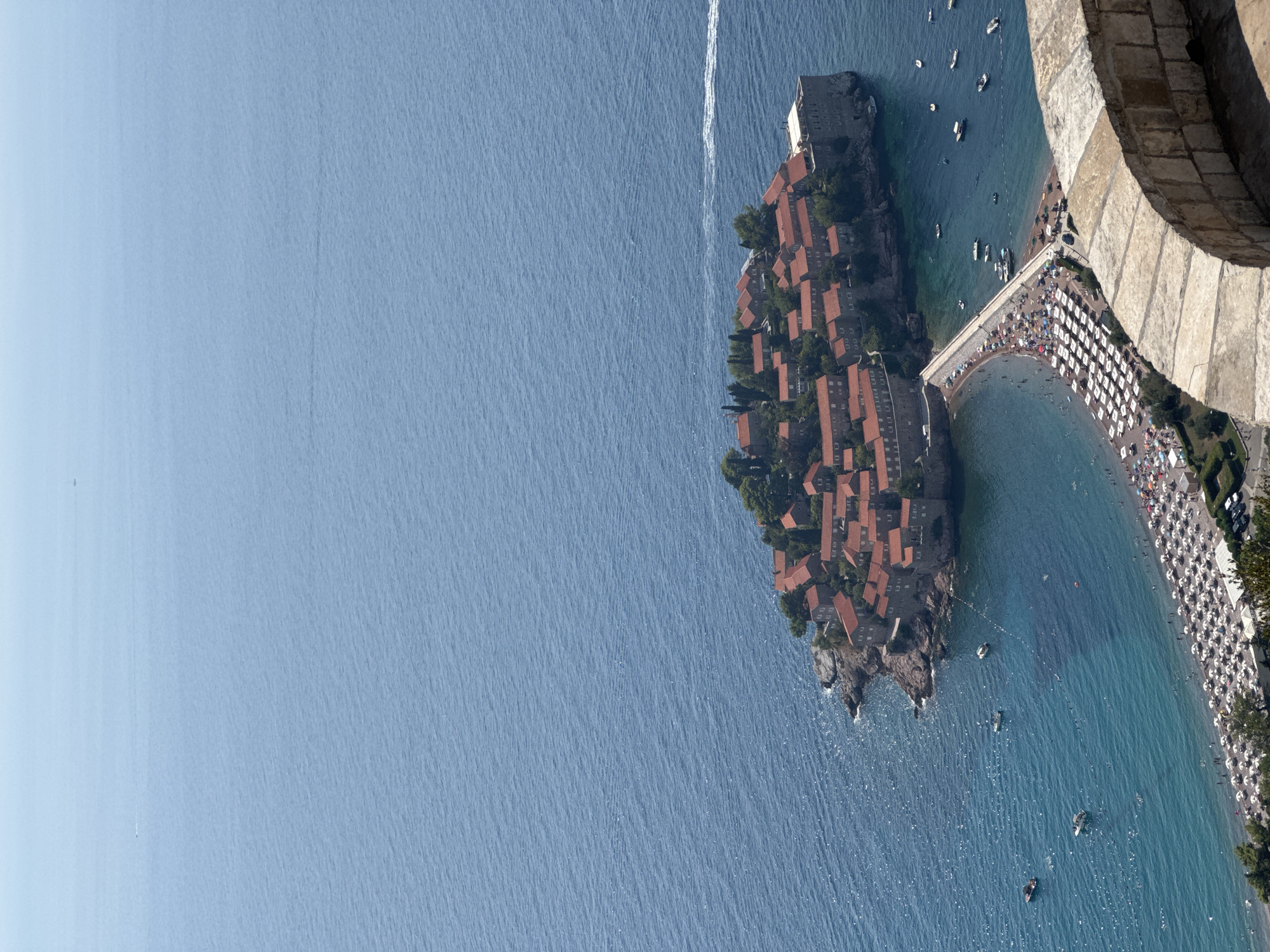 An aerial, high-angle, full outdoor shot depicts the city of Sveti Stefan, Montenegro, with a focus on the island and its connecting causeway. The island is a cluster of red-roofed buildings, densely packed and surrounded by trees. The water is a clear turquoise, with several boats anchored or moving in its vicinity. A causeway made of paving stones connects the island to a sandy beach lined with white umbrellas and lounge chairs, where many people are visible, some swimming. The sea extends to the horizon, a vast expanse of blue. The scene is bathed in sunlight, casting sharp shadows. In the foreground, a curved stone wall with a rough texture is visible.