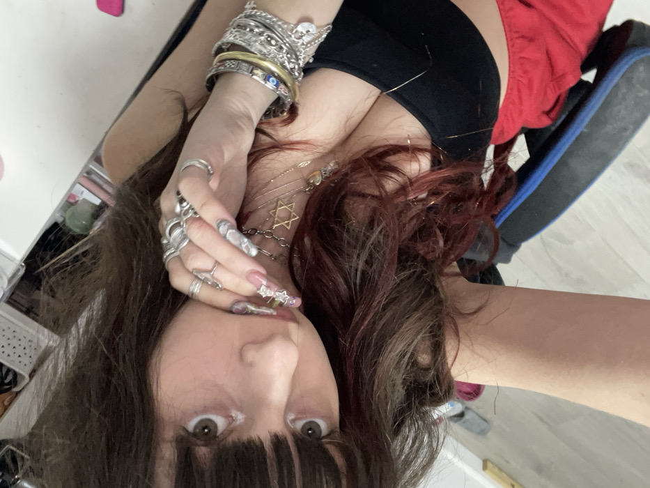 A young woman gazes upward. She has long brown hair with red highlights and full bangs. She wears many rings on one hand with a stack of bracelets on her wrist. Several necklaces adorn her neck. She wears a black top and red bottoms. She appears to be sitting in a chair. The background is a combination of a wall and floor.