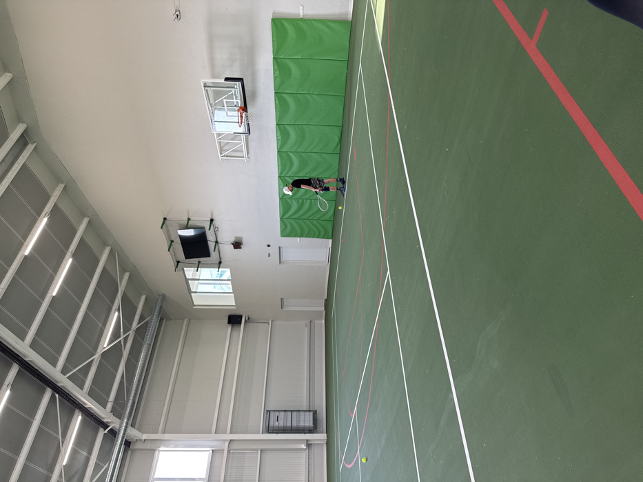 This image depicts an indoor sports hall located in the city of Gorna Mitropolia, Bulgaria. The facility features a dark green court with prominent white and red lines, delineating areas for tennis and potentially other court sports. A single adult male is positioned on the court, holding a tennis racket with the head oriented towards the ground. He is dressed in a white baseball cap, a dark short-sleeved shirt, dark patterned shorts, and athletic shoes. A knee brace is visible on his right knee. Two yellow tennis balls are present on the court: one near his right foot and another further into the mid-court area, to the left of the center line.
The hall has high, light-colored walls and a sloped ceiling with exposed white metal trusses and multiple linear fluorescent light fixtures. Natural light enters through large rectangular windows on the left wall and smaller elevated windows on the far wall. A large black flat-screen monitor, enclosed within a protective net, is mounted high on the far wall. Below it, two white doors are visible. To the right of the doors, a basketball hoop with a transparent backboard is installed. Below the basketball hoop and extending across the far wall, a series of light green padded panels are affixed. A ventilation duct runs along the upper left wall, where a black speaker is also mounted. A large grey metal utility or storage unit is visible against the lower section of the left wall.