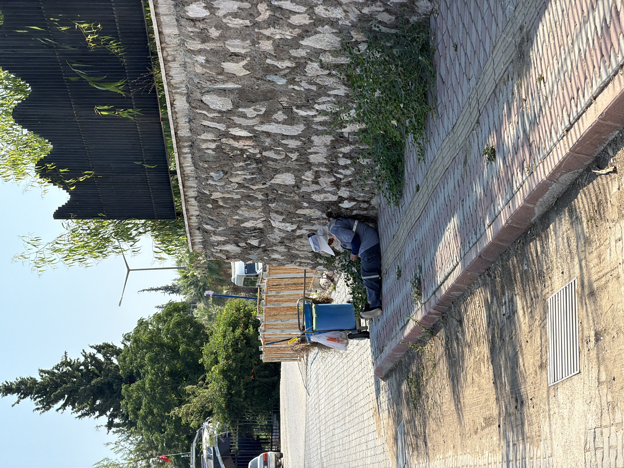 In Kemer, Turkey, a person in a light-colored hat and grey work uniform is resting against a stone wall on a paved sidewalk. The individual is seated with their legs extended and their head tilted down, appearing to be asleep or taking a break. Beside them is a blue trash cart with a bag and cleaning tools, including a broom. The sidewalk is bordered by a dirt path and a raised curb. The background features a stone wall, green foliage, a tall tree, a street light, and a black metal fence with a decorative wavy top. Buildings and vehicles are visible further in the distance. The scene is set under a clear blue sky on a sunny day, with shadows cast by trees and the fence.