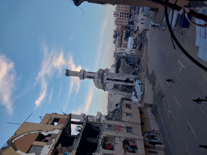 The image depicts a heavily damaged urban landscape, viewed from an elevated position, likely from a building or upper floor. The overall scene conveys a sense of desolation and destruction.
In the center-left, a prominent minaret, part of a mosque, stands tall against the sky. The minaret itself shows signs of damage, with sections appearing eroded or broken. The mosque structure connected to it also exhibits significant damage, with large parts of its walls and roof missing or collapsed, revealing interior debris.
The foreground and midground are dominated by ruined buildings. On the left side, a multi-story building is extensively damaged, with large sections of its facade torn away, exposing internal rooms and structural elements. Debris, including twisted metal and shattered concrete, is visible both within the building and scattered around its base. Similar damage is apparent on other buildings throughout the scene, with many structures appearing as hollow shells or piles of rubble.
A paved road runs through the middle of the image, showing clear lane markings, including a dashed white line and a white arrow painted on the asphalt, indicating a direction of travel. Several figures of people are visible on the road and sidewalks, walking or standing, navigating the debris-strewn environment. Their presence suggests that despite the destruction, the area is still inhabited or traversed.
The background shows more urban structures, some appearing intact while others contribute to the overall impression of a city scarred by conflict or disaster. On the right side, there are hints of makeshift shelters or tents, possibly indicating displaced populations or temporary arrangements. Dark cables or wires are visible in the extreme foreground, hanging and crisscrossing the upper right portion of the frame, possibly remnants of electrical lines or communication infrastructure.
The sky above is a clear blue with scattered, wispy white clouds, suggesting fair weather. The lighting indicates either early morning or late afternoon, casting soft shadows and highlighting the texture of the damaged buildings. No clearly legible text is visible on any signs or banners due to distance and resolution. The scene evokes a strong sense of loss and the aftermath of a devastating event.