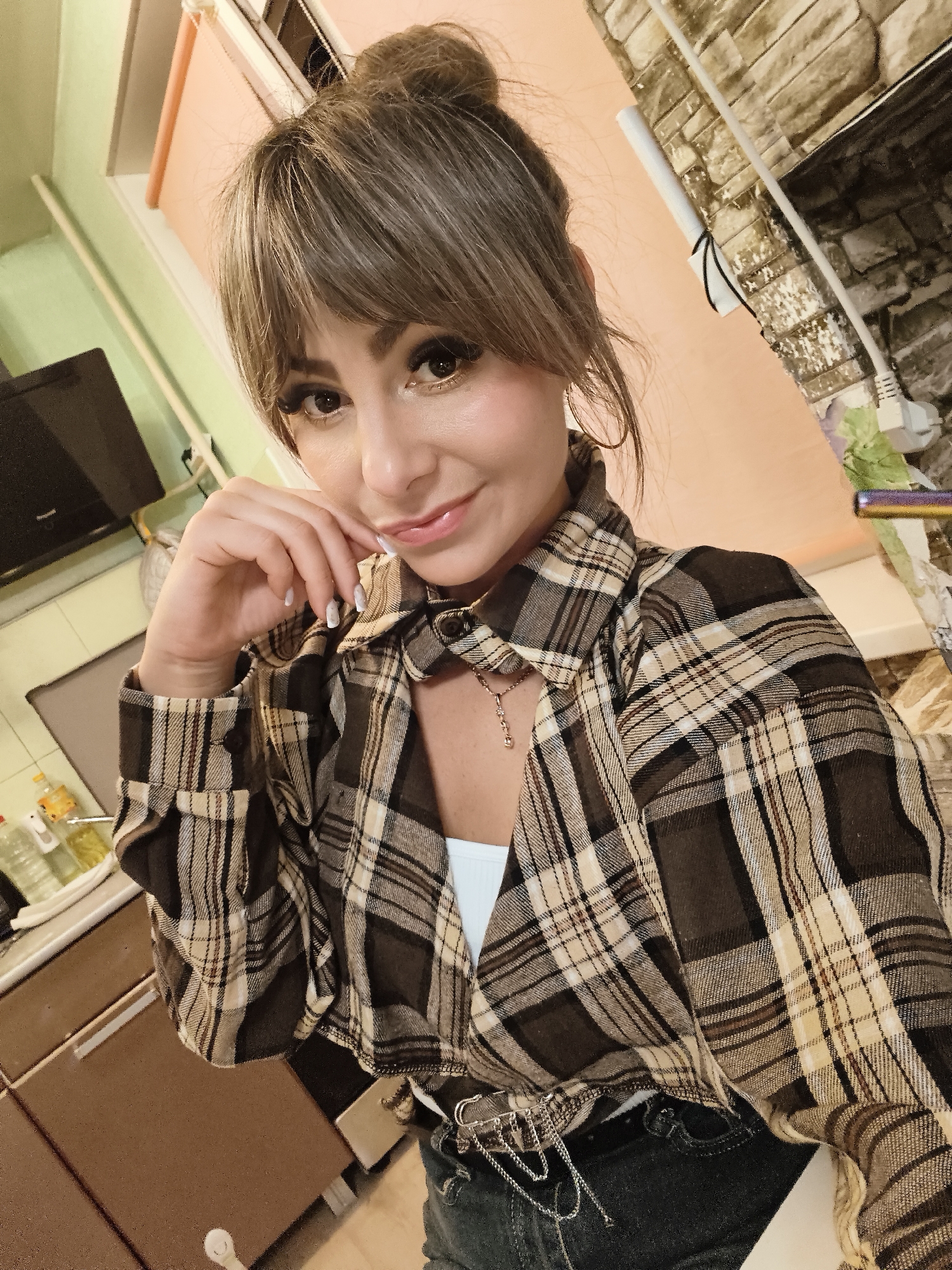 A woman is posing for a selfie indoors. She has brown hair pulled back in a messy bun with bangs, brown eyes, and wears makeup including mascara and eyeliner. She is wearing a brown and beige plaid shirt over a white top. A silver chain belt is visible around her waist. She has a gold necklace and hoop earrings. Her left hand is resting against her face. The background includes a kitchen area with cabinets, a TV, a window with a peach-colored blind, and a wall with a textured stone-like pattern. The location appears to be a home in Chaikovsky, Russia.