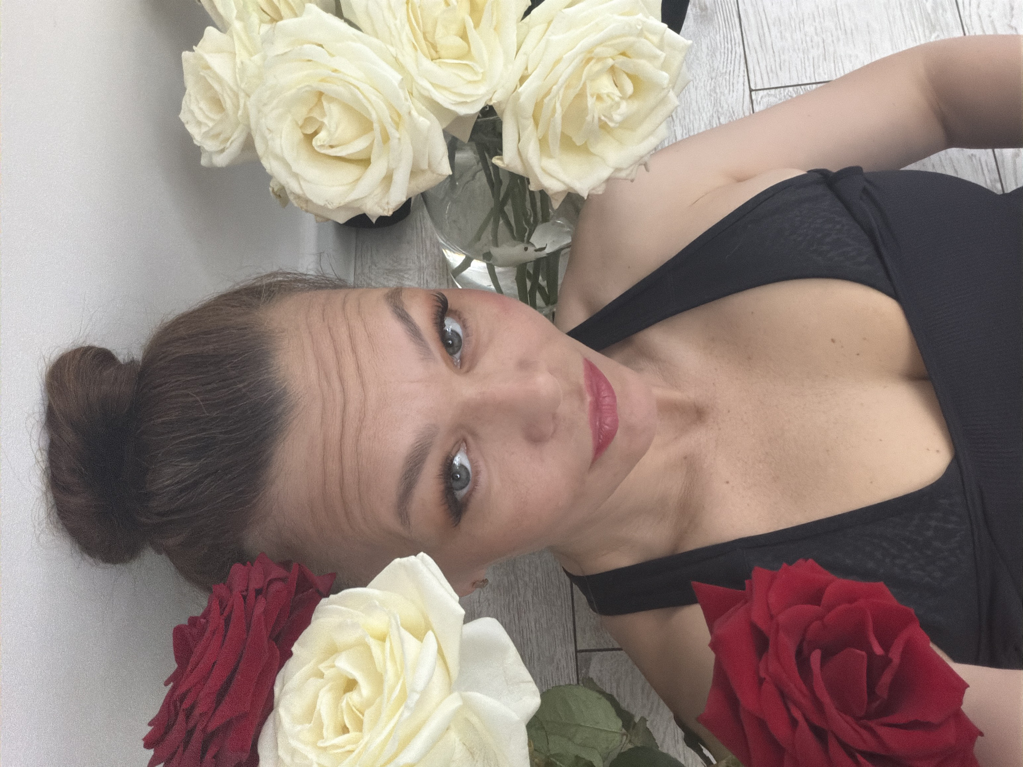 A woman lies on a wooden floor in a location identified as Chaikovsky, Russia. She is wearing a black top and is surrounded by roses. There is a vase of white roses near her head, and individual red and white roses near her shoulders. Her hair is in a bun, and she is looking up at the camera with a slight smile.