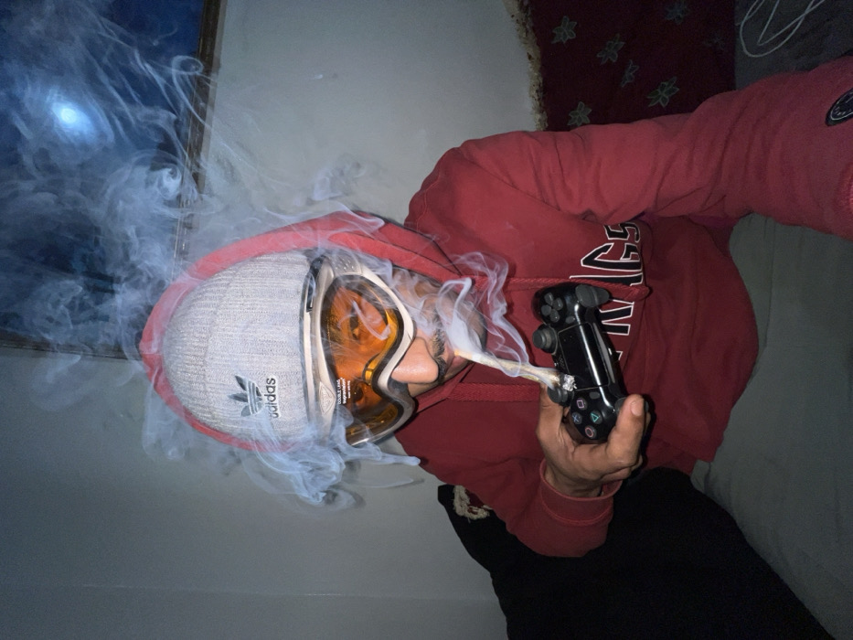 An individual is shown indoors, partially obscured by a dense cloud of white smoke. The person wears a grey Adidas beanie, orange-tinted ski goggles, and a red hooded sweatshirt with the hood pulled up. A dark-colored PlayStation gaming controller is held in the right hand. A lit, hand-rolled cigarette-like object is inserted into the controller's top-right joystick area, actively burning and producing the surrounding smoke. The individual has a mustache and is looking forward. The background consists of a plain, light-colored wall. A framed item on the upper right wall reflects a bright, circular light source. A dark sofa or cushion is visible on the left, and a patterned rug or textile, exhibiting motifs consistent with Moroccan interior design, covers the floor area to the right. The scene takes place within a residential room; no specific landmarks of Tanger, Morocco, are visible to confirm the city location, although the interior decoration aligns with Moroccan aesthetics.