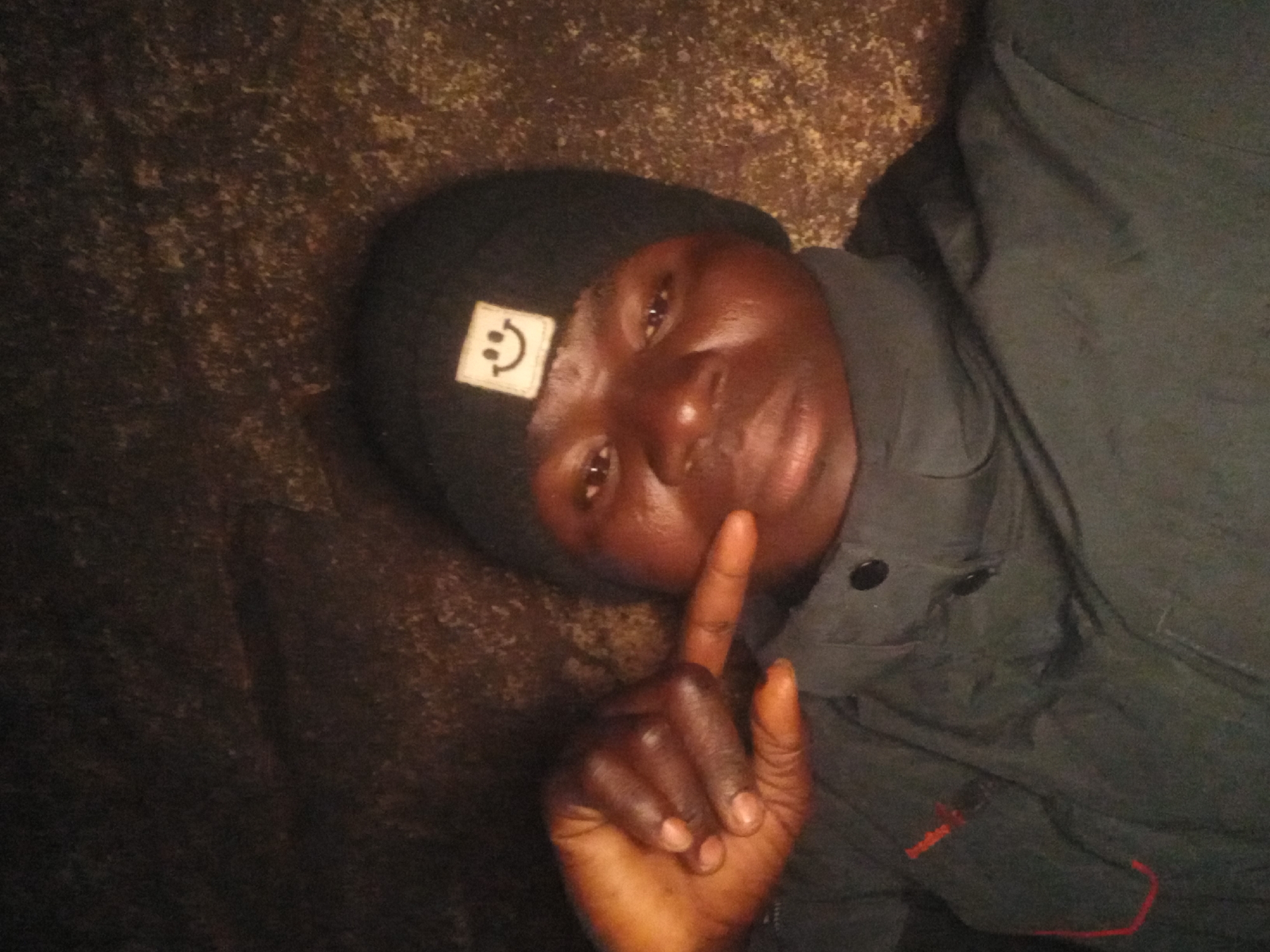 The image is a selfie taken by a person wearing a black beanie with a white smiley face emblem and a dark jacket. The person is holding up their right hand, with fingers extended and pointing towards the left side of the frame. The background appears to be a rough, textured surface, possibly rock or a wall, with mottled brown and dark tones. The lighting is dim, creating shadows and a somewhat grainy quality to the image. The person's face is partially illuminated. The location is identified as Kwang, Nigeria.