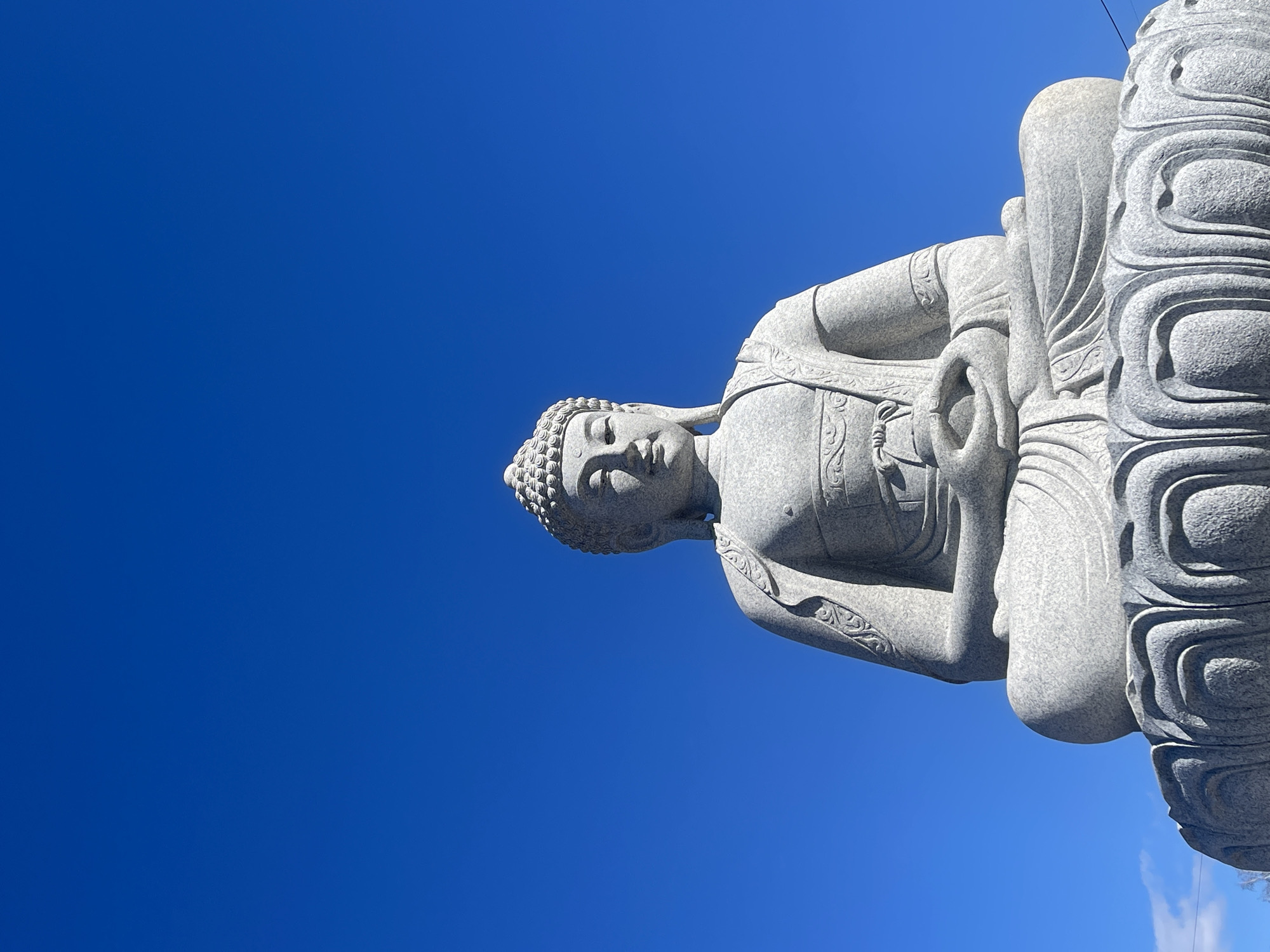 The image shows a statue of Buddha sitting in a lotus position against a clear blue sky. The statue appears to be made of gray stone. It is located in Krasnoyarsk, Russia.