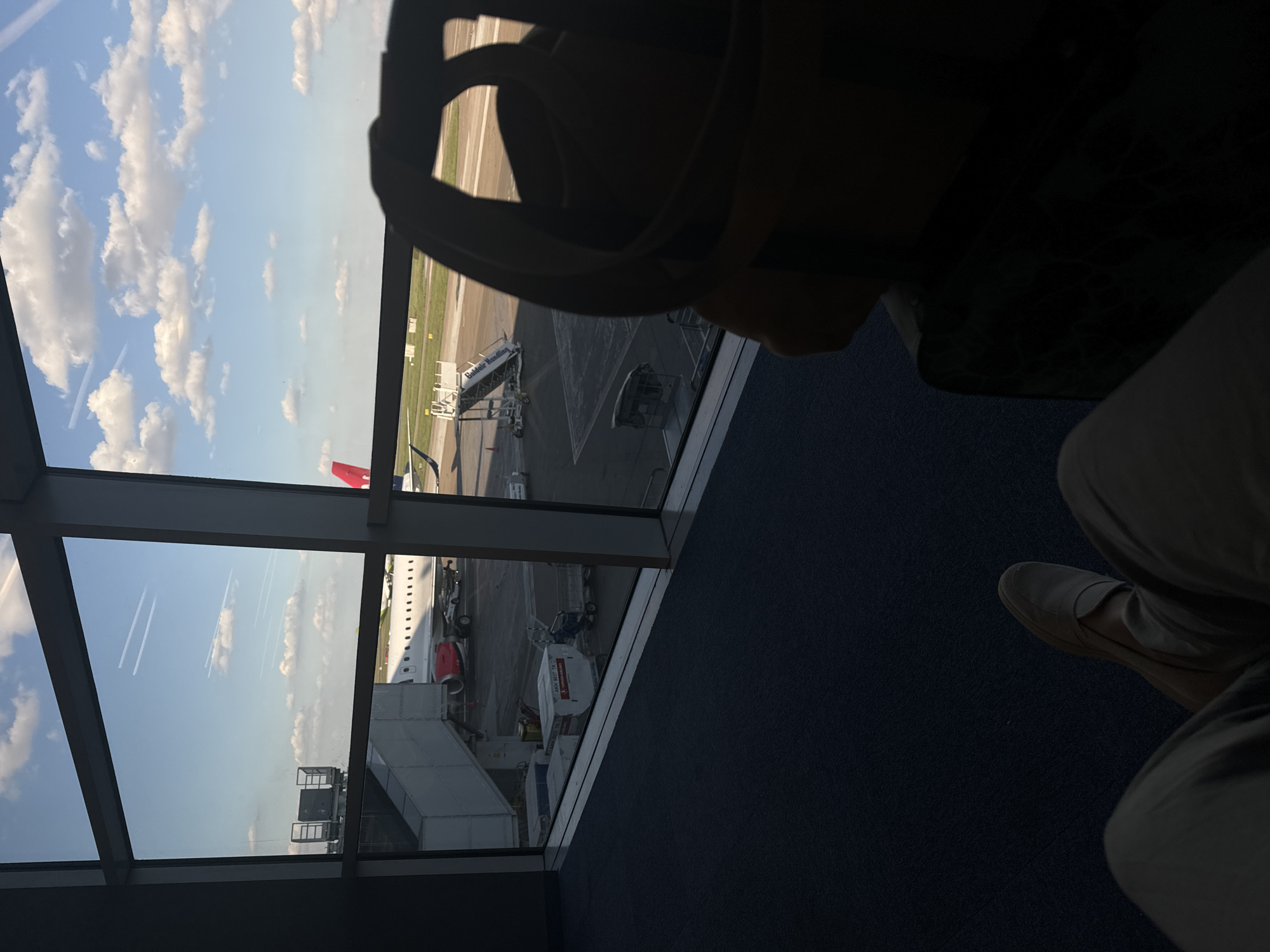 The image is taken from inside an airport, likely in Surcin Urban Municipality, Serbia, given the plane markings and airport infrastructure. The view is out a window, showing an airplane docked at a gate. There are jetways, baggage handling equipment, and other airport vehicles visible. In the foreground, a person is seated, wearing what appears to be a headset or headphone-like device. The person is partially visible, and their leg with a shoe is in the frame. The window reflects the sky, with clouds.