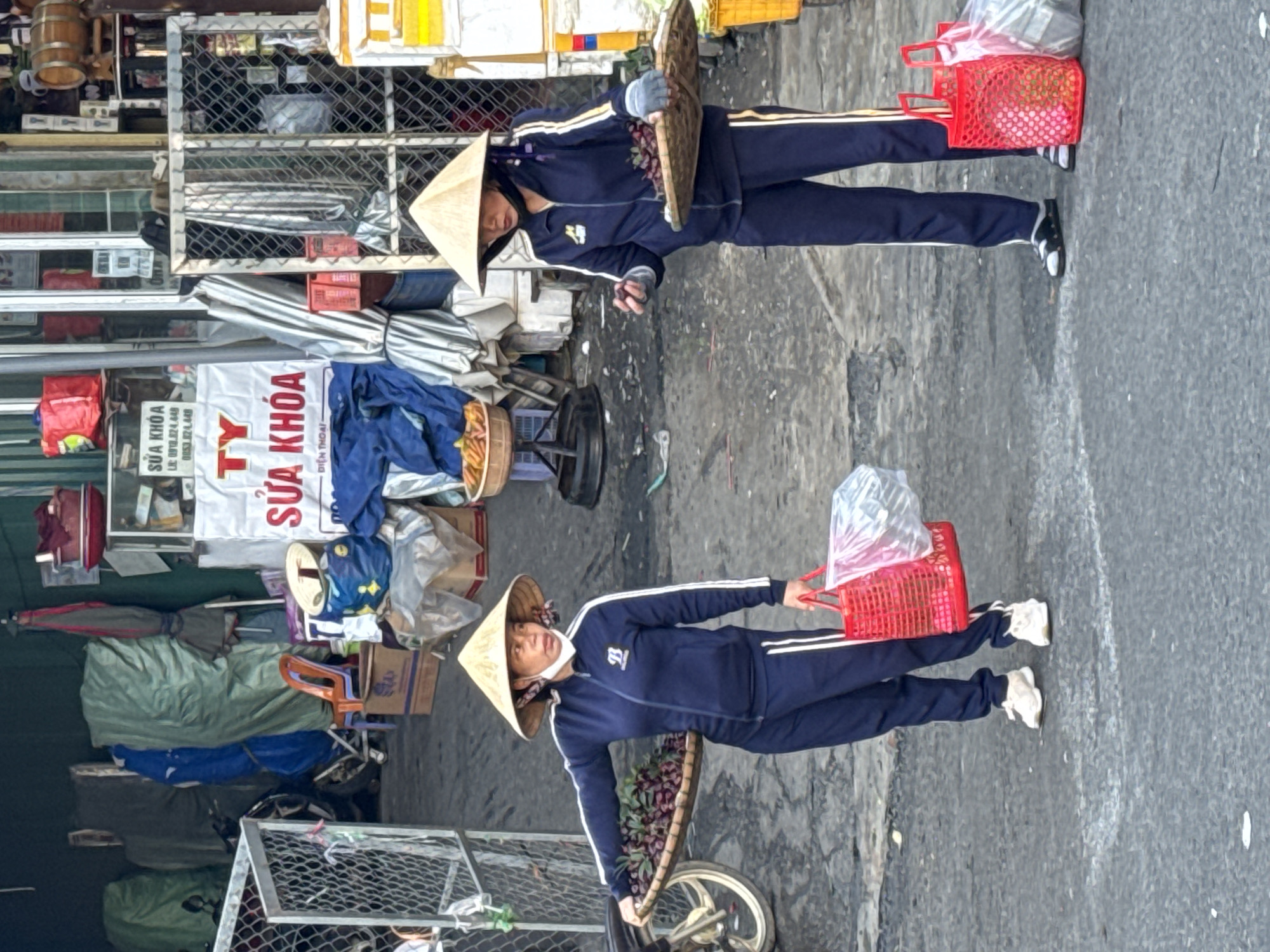 Two women wearing conical hats and matching dark blue tracksuits stand on a paved surface in what appears to be an outdoor market or street stall area. The woman on the left holds a flat, woven tray filled with dark purple fruit, possibly grapes or plums, in her left arm and carries a red plastic basket in her right hand. She wears white sneakers. The woman on the right holds a similar woven tray of fruit with her left hand and has a red plastic basket at her side, containing plastic bags. She wears black slip-on sandals. Both women are wearing light-colored face coverings. In the background, there are various market stalls, including displays of goods, metal cages, and what looks like a motorcycle parked behind a fence. The environment suggests a busy, urban setting in Da Lat, Vietnam, likely a local market where produce is sold.