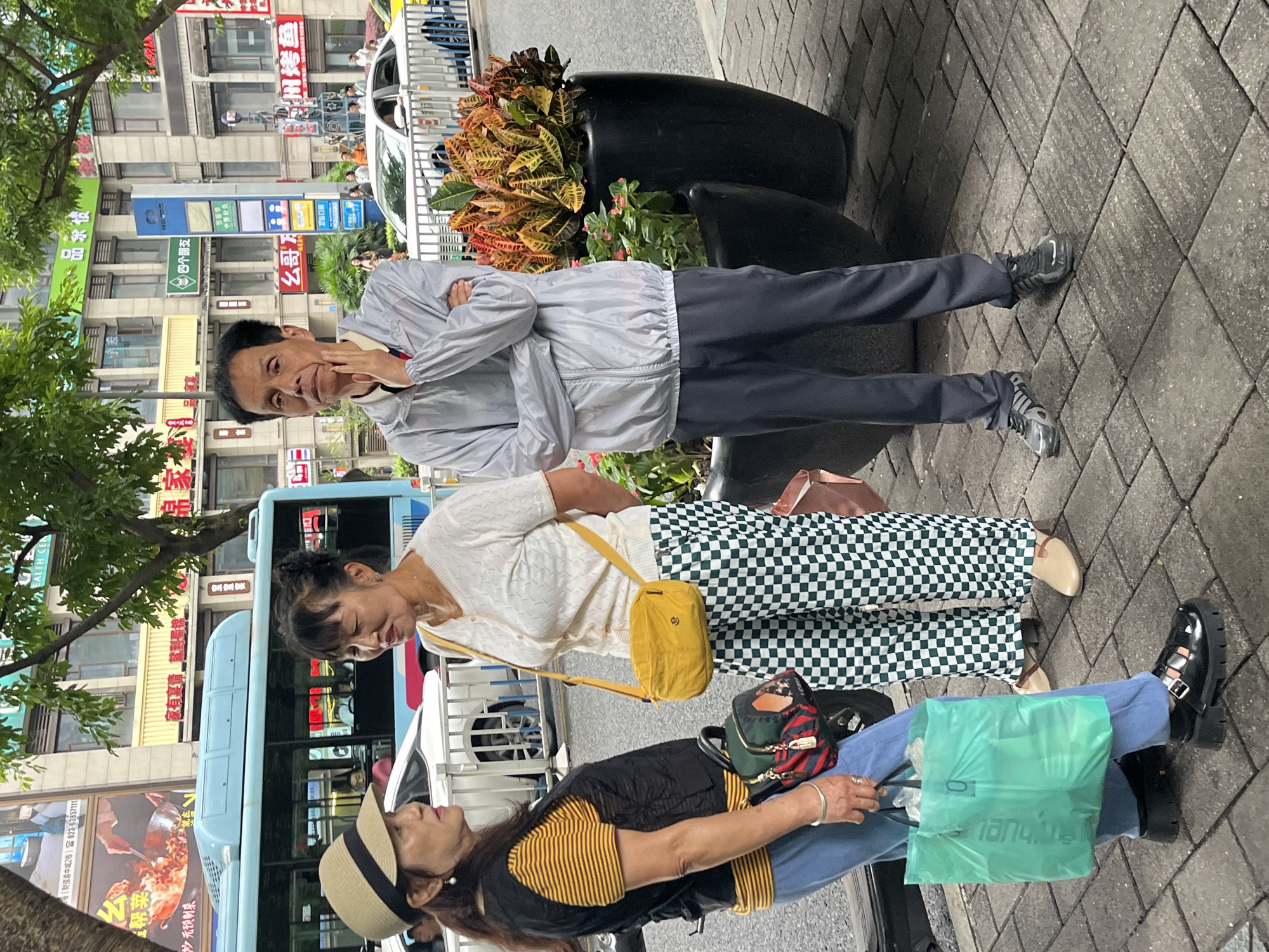 The image depicts a street scene in the Yuzhong District of China, where three people are standing on a tiled sidewalk. On the left, a woman wearing a straw hat, a yellow and black striped shirt, and a black vest is holding a green shopping bag. She is looking towards another woman standing to her right. This second woman is wearing a white knitted shirt and green and white checkered pants, and she has a yellow cross-body bag. She is looking down. In the center of the image, a man in a light grey windbreaker and dark pants stands with his arms crossed, his hand resting on his chin, looking towards the viewer. Behind the people, a light blue bus is visible, as well as buildings and trees. Large black planters with colorful plants are placed near the sidewalk.