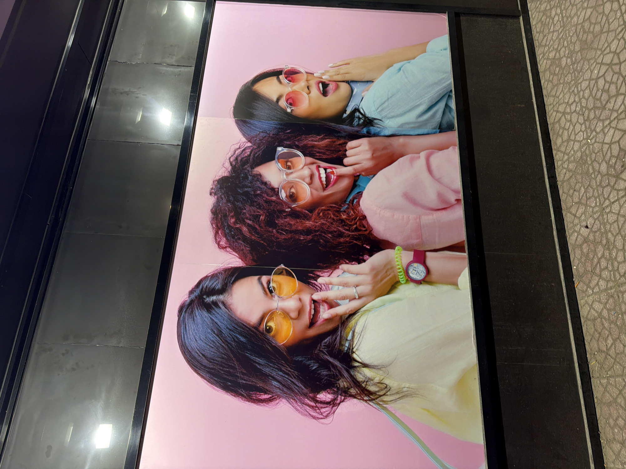 A wide, horizontal billboard-style advertisement is displayed on a pink background. The ad features three young women, each wearing large, tinted sunglasses. The woman on the left has dark hair and is wearing a pale yellow top, orange sunglasses, and a watch with a pink band and green strap. The woman in the middle has curly, dark hair and is wearing a pink top and clear-rimmed glasses with orange tint. The woman on the right has dark hair and is wearing a light blue top and clear-rimmed glasses with pink tint. All three women have their mouths open, appearing surprised or excited, and are looking towards the viewer. The advertisement is framed by a black border, and the bottom of the frame shows a cobblestone pavement. The scene appears to be an outdoor urban setting, likely a street or a public space in Almaty, Kazakhstan.