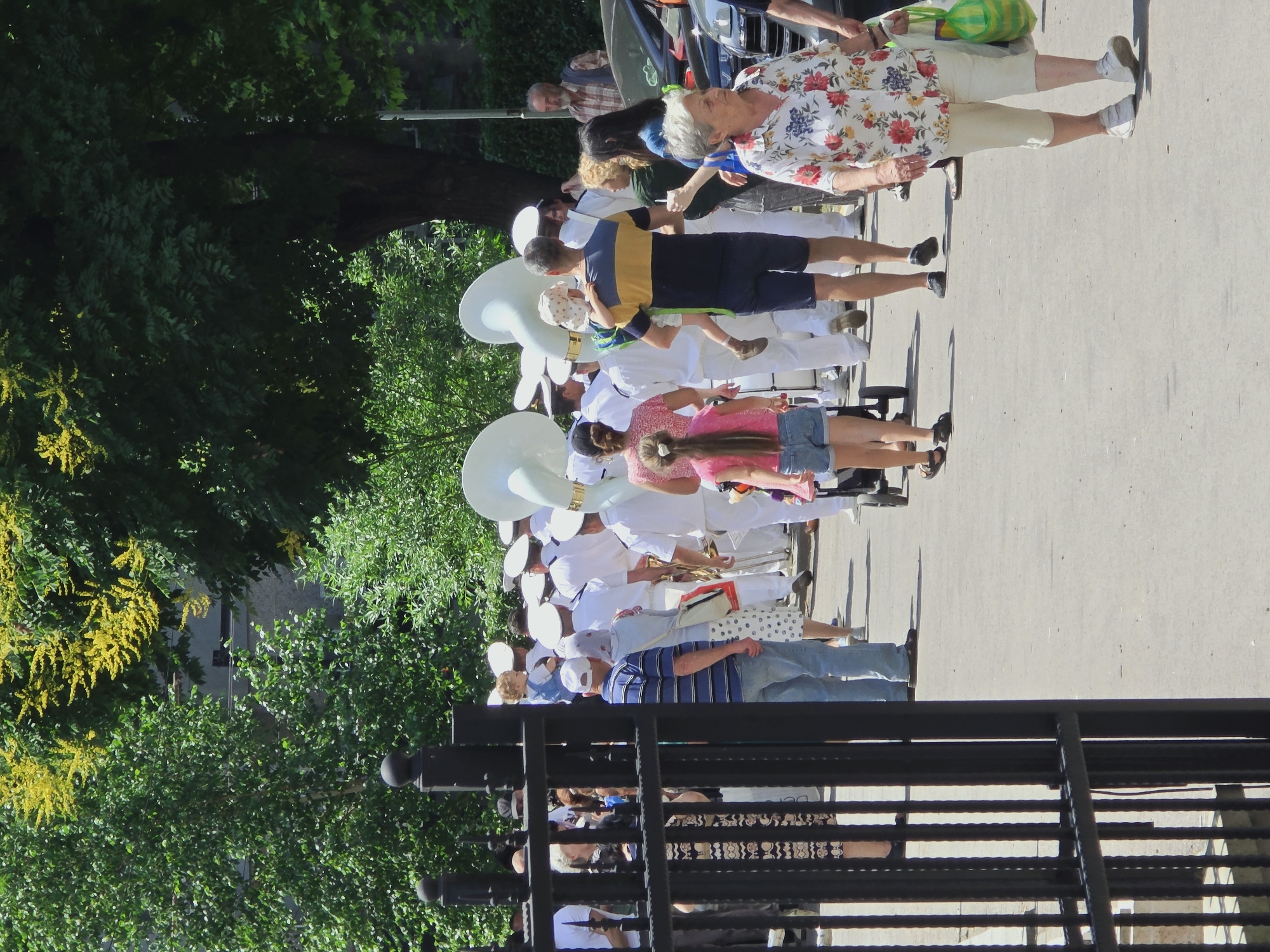 The image depicts a street scene in Varna, Bulgaria, possibly during a parade or event. In the foreground, a black metal fence is visible on the left, obscuring part of the scene. Several people are walking on a paved surface. A man in a blue and white striped shirt and blue jeans walks away from the camera, while a woman in a white shirt with a floral pattern and light-colored pants walks towards the camera on the right, carrying a green plastic bag. Behind them, a group of individuals, some in white naval uniforms with white hats, are walking. Two large, white brass instruments, possibly tubas, are being carried or are part of the procession. In the background, trees with green foliage and some yellow flowers are present, suggesting it is springtime or summer. The overall atmosphere appears to be daytime and outdoors.