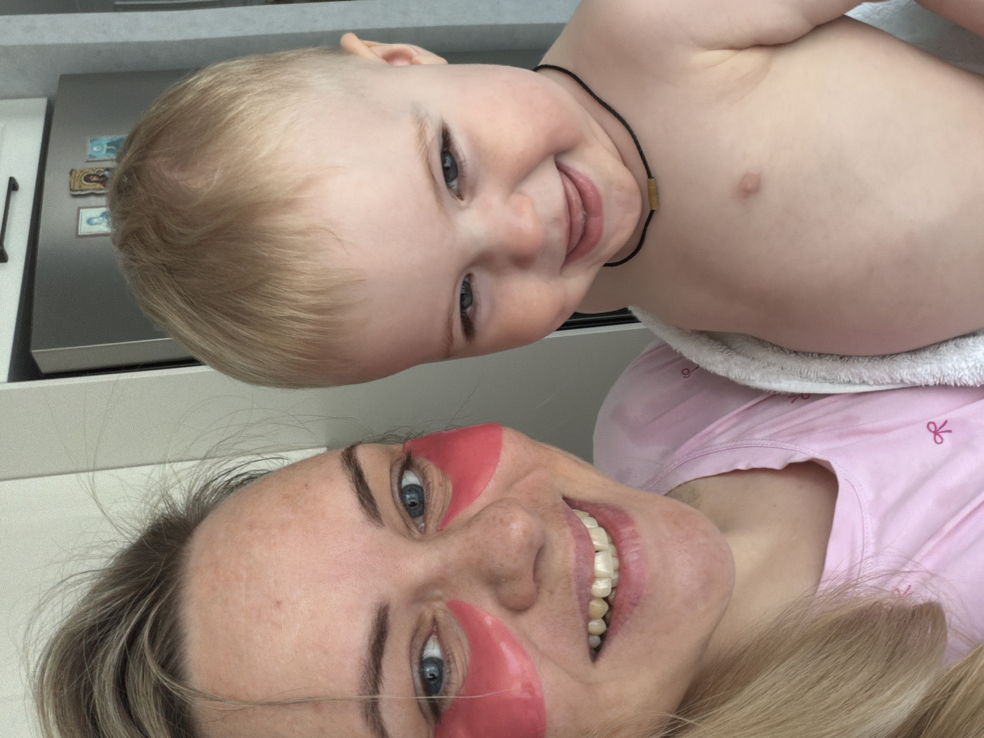 A selfie depicts a woman with pink under-eye patches and blonde hair lying down, with a fair-skinned baby in the upper portion of the frame. The baby has blonde hair, blue eyes, and is wearing a black cord necklace with a gold accent. The woman is wearing a pink shirt. The background includes a silver refrigerator with magnets on it, and white cabinetry. The image does not contain information to determine if it was taken in Votkinsk, Russia.
