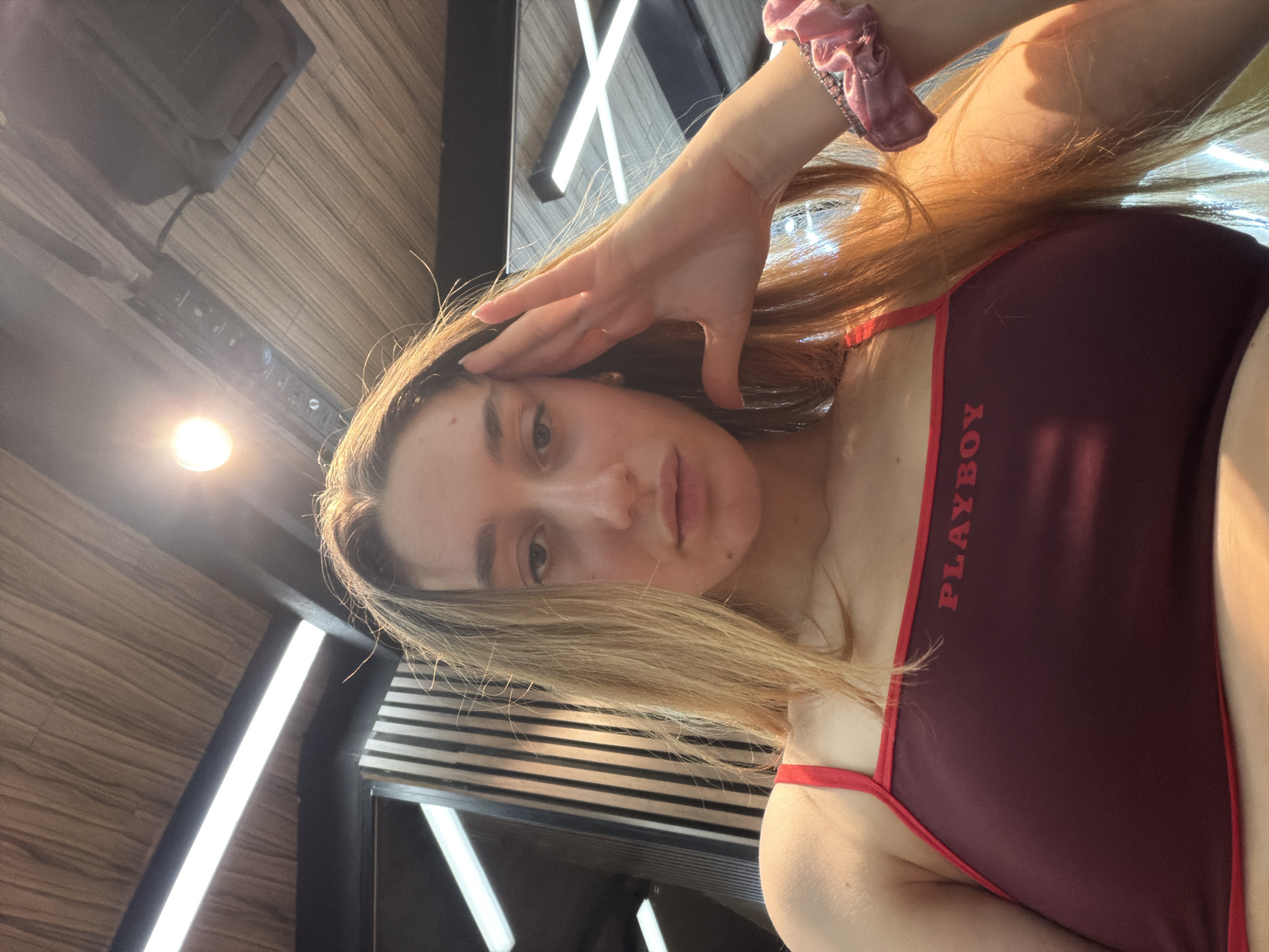 A woman is facing the camera with her hand on her forehead. She is wearing a maroon top with red trim, the word "PLAYBOY" printed vertically on it. Her hair is light brown. She has a pink scrunchie in her hair. The background consists of a wooden panel ceiling, an array of linear light fixtures and horizontal blinds. There is no indication that this photo was taken in Sofia, Bulgaria.