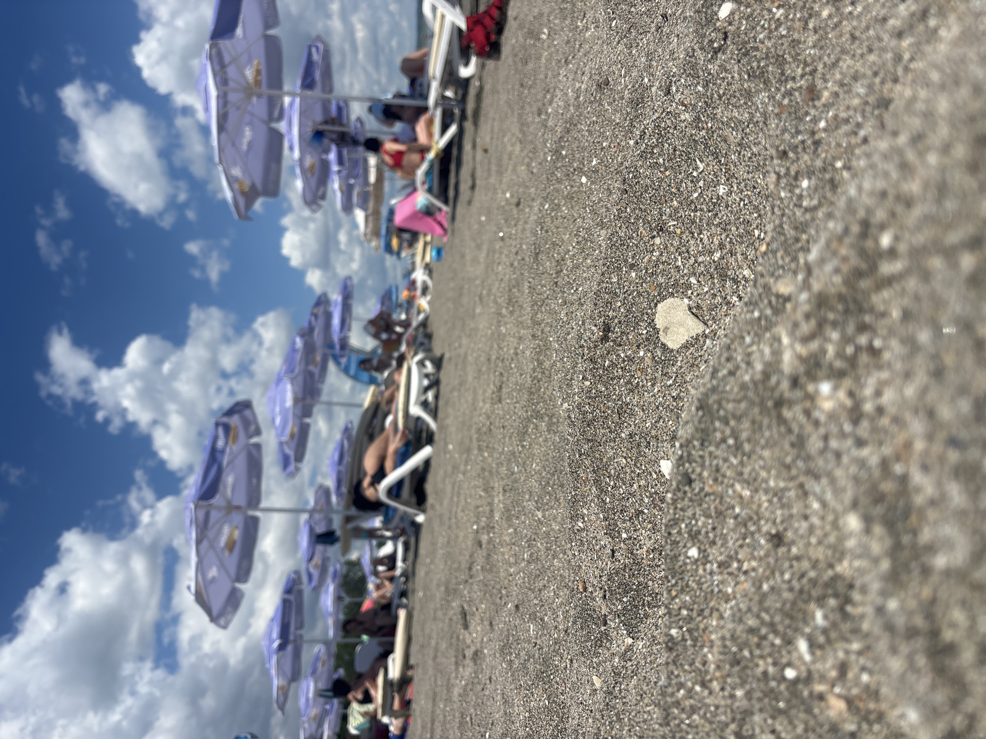 In an outdoor scene, the foreground is dominated by the texture of dark sand, with a single small, heart-shaped shell clearly visible. In the mid-ground, several people recline on lounge chairs under an array of purple beach umbrellas. Some individuals are partially visible, engaged in leisure activities. The background features more beach umbrellas and a hazy view of the sea or body of water. The sky above is a vibrant blue, dotted with scattered white clouds, suggesting a sunny day.