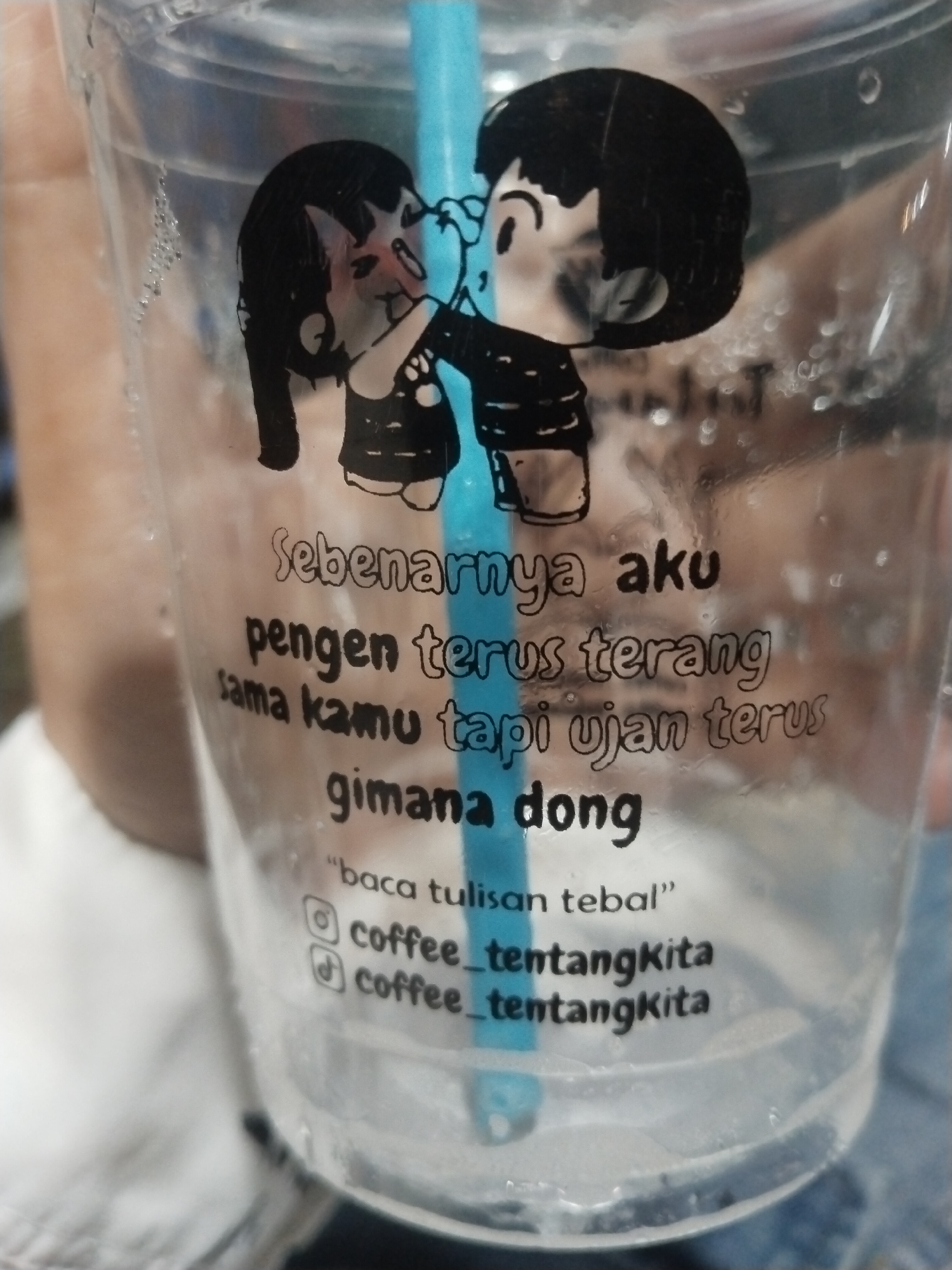 This media file is a close-up shot of a clear plastic cup with text and an illustration printed on it. The cup is filled with a clear liquid, likely water or a beverage, and a bright blue straw is visible inside. The illustration depicts two cartoon figures, a boy and a girl, in a close embrace with the boy whispering into the girl's ear.
Printed on the cup in black, handwritten-style text is a romantic and slightly melancholic message in Indonesian. It reads: "Sebenarnya aku pengen terus terang sama kamu tapi ujan terus gimana dong" which translates to "Actually, I want to be upfront with you, but it keeps raining, what should I do?". Below this, in smaller text, is a directive: "baca tulisan tebal" meaning "read the bold text". Finally, two social media handles are listed, both for Instagram: "@coffee_tentangkita" and "☑ coffee_tentangkita". The checkmark next to the second handle indicates a verified account.
The overall mood evoked is one of gentle longing and perhaps a touch of playful frustration. The presence of condensation on the outside of the cup suggests it might be a cold drink, and the mention of rain in the text could imply a cloudy or wet day, or it could be a metaphorical expression of emotional state. The setting appears to be casual, given the nature of the cup and its inscription, possibly a cafe or a place where take-away drinks are served in Lhokseumawe, Indonesia. The lighting is soft, suggesting it might be daytime, but the exact time of day is not definitively clear.