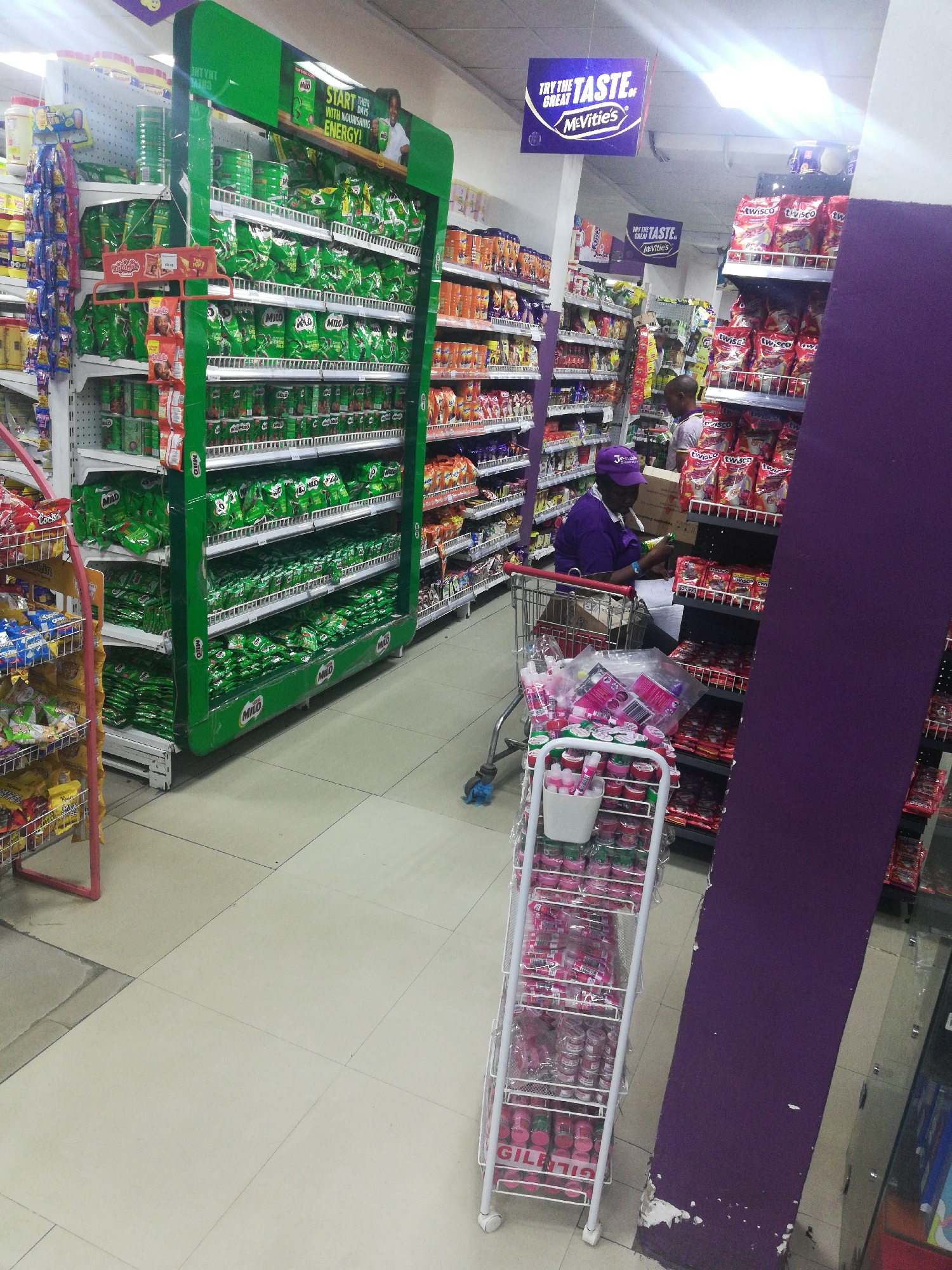 The image captures the interior of a supermarket in Lagos, Nigeria. Aisles filled with products such as Milo are visible. A person wearing a purple shirt and cap is examining items in their shopping cart. Another individual is visible further down the aisle. The scene is lit with fluorescent lighting. A cart full of small items, including what appear to be skincare or cosmetic products, is located in the lower foreground. Signage promoting "McVitie's" is suspended from the ceiling. A prominent purple pillar is in the right of the image.