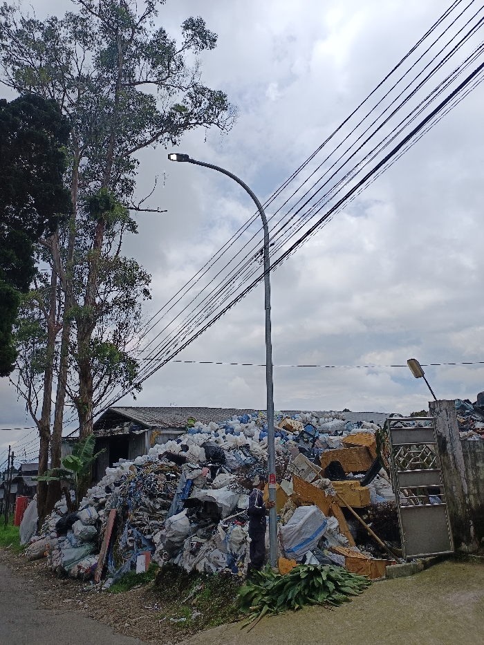 A massive pile of mixed trash dominates the foreground, partially obscuring a building with a grey roof. A man stands near a light pole amidst the waste. Power lines stretch diagonally across the overcast sky. Vegetation lines the roadside near the trash. A partial fence and gate are visible on the right side of the waste pile.