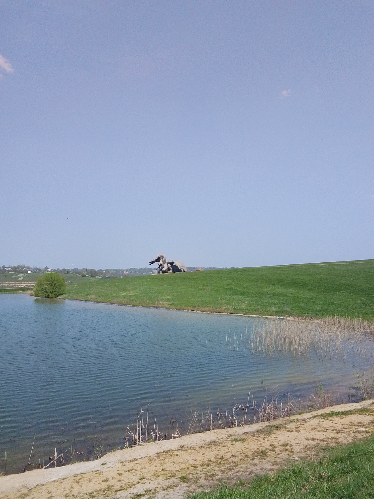 The image captures a tranquil scene featuring a body of water, likely a pond or small lake, with gently rippling water. On the opposite bank, a grassy hill rises, topped by what appears to be a sculpture or artistic installation. The sculpture has a grey or earthen tone, contrasting with the bright green grass. A small, round tree stands near the water's edge, adding to the natural setting. The sky is a clear, light blue, with a few wispy clouds visible. The foreground consists of a sandy or earthen bank with sparse vegetation. The overall impression is a peaceful, natural landscape with an artistic element.
Based on the information provided the photograph was taken in the city of Каменка, Russia.
