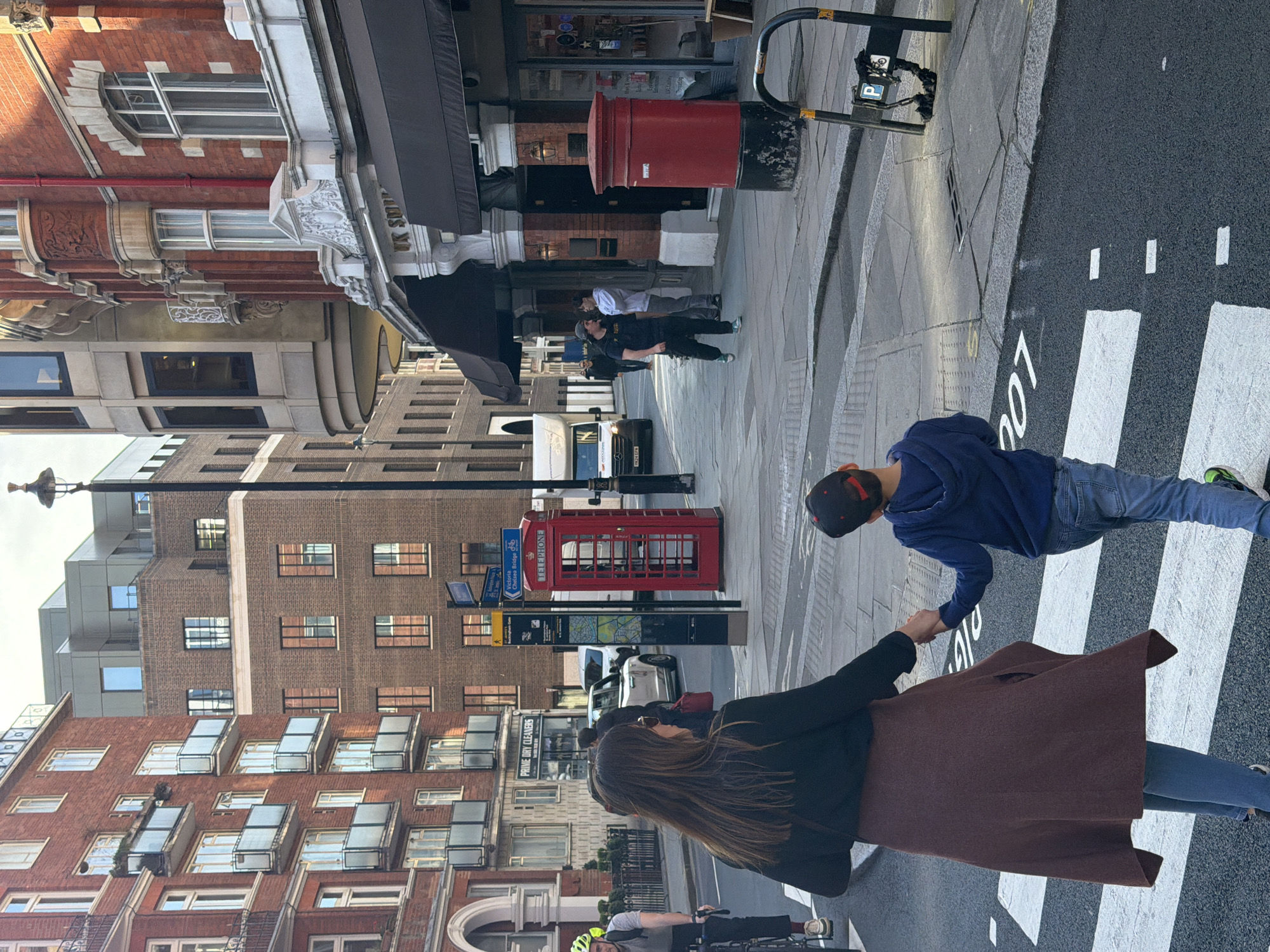 In the city of London, United Kingdom, a medium shot captures a mother and son walking across a pedestrian crosswalk. The mother, with long brown hair and sunglasses, wears a brown coat over a black jacket and blue jeans. She holds the hand of her son, who is wearing a blue sweatshirt, jeans, and a black baseball cap. They are mid-stride on the zebra crossing.
Behind them, a classic red telephone box stands on the sidewalk. Further down the street, a red post box is visible. The street is lined with brick buildings of varying heights and architectural styles. A bicyclist passes in the lower part of the frame, and cars are visible parked along the road. The environment suggests a bustling urban setting in London.