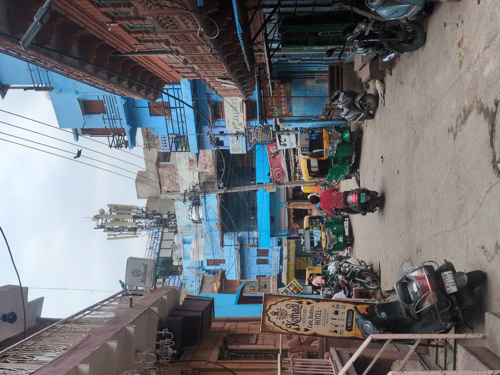 In Jodhpur, India, a street scene unfolds with a narrow lane flanked by buildings of varying architectural styles and colors. The buildings on the left are a muted pink and cream, featuring a balcony with decorative railings. To the right, a vibrant blue building stands prominently, with wooden balconies and windows. Further to the right, a building with a rustic, weathered brick facade displays ornate wooden latticework balconies.
The street is a mix of paved and uneven surfaces, with visible debris and stains on the ground. Several vehicles are parked or in motion, including a green and yellow auto-rickshaw in the center of the frame, and multiple scooters and motorcycles, some parked along the side and others being ridden. A person in a red shirt is riding a scooter, heading away from the camera. Another person in a light pink shirt is standing near an auto-rickshaw. A dog is lying down in the middle of the street.
Prominent signage includes a tall, vertical sign for "Kothadi Cafe, Restro & Hotel," featuring traditional Indian motifs and a phone number. Other signs, in both English and Hindi, are visible on the buildings, indicating various businesses and establishments. Overhead, a complex network of electrical wires and a cellular tower with multiple antennae dominate the sky, which is overcast and appears to be a pale gray. The overall impression is one of a busy, lived-in urban environment.