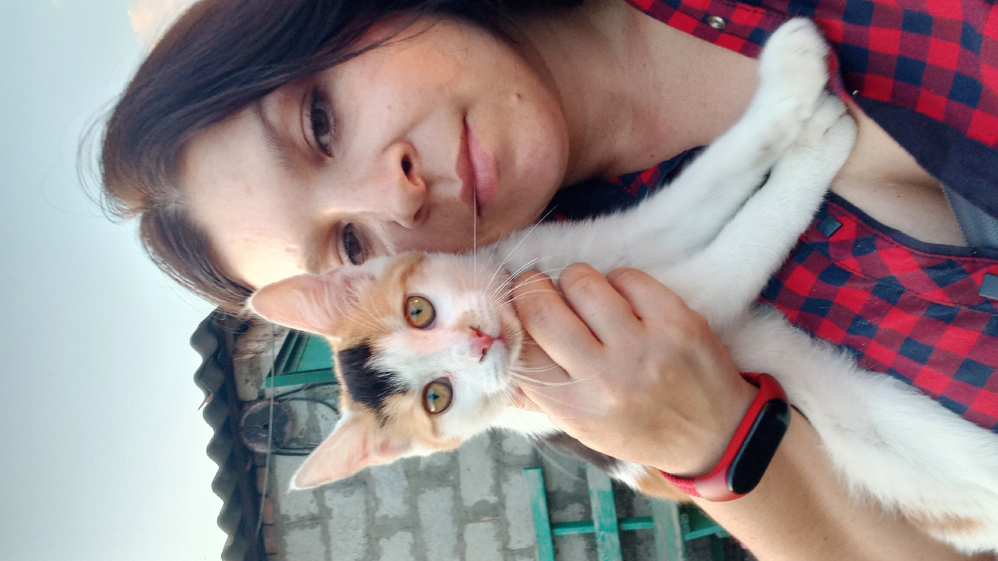 In a close-up selfie taken outdoors, a young woman with dark hair and brown eyes holds a calico kitten. She is wearing a red and black plaid shirt. The kitten, with white, orange, and black fur and striking yellow eyes, is nestled against her chest and held by her left hand. A thin strand of what appears to be a cat's whisker or hair rests on her upper lip. On her left wrist, she wears a red fitness tracker. The background suggests a rustic setting with a corrugated metal roof of a building and a grey brick wall, along with a glimpse of a green bench. The sky above is a pale blue, indicating daytime. The scene is captured in Klyntsi, Ukraine.