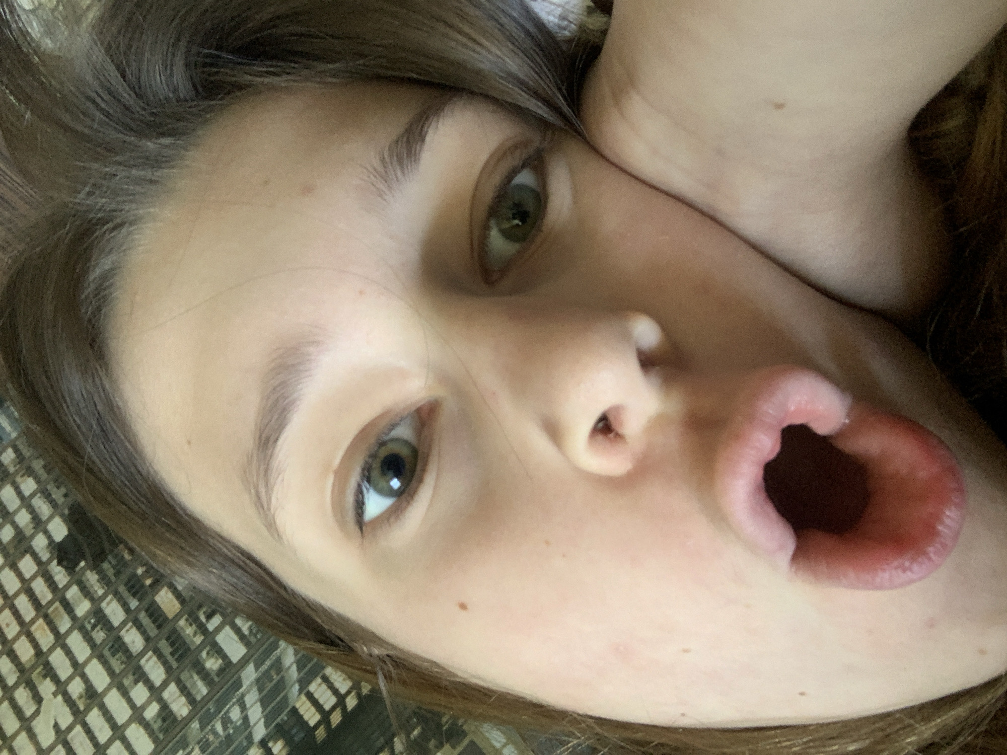 A close-up, angled selfie of a Caucasian female with light brown hair and green eyes. Her mouth is open wide. She is lying on a surface with a woven texture. The location is not discernible from the image.