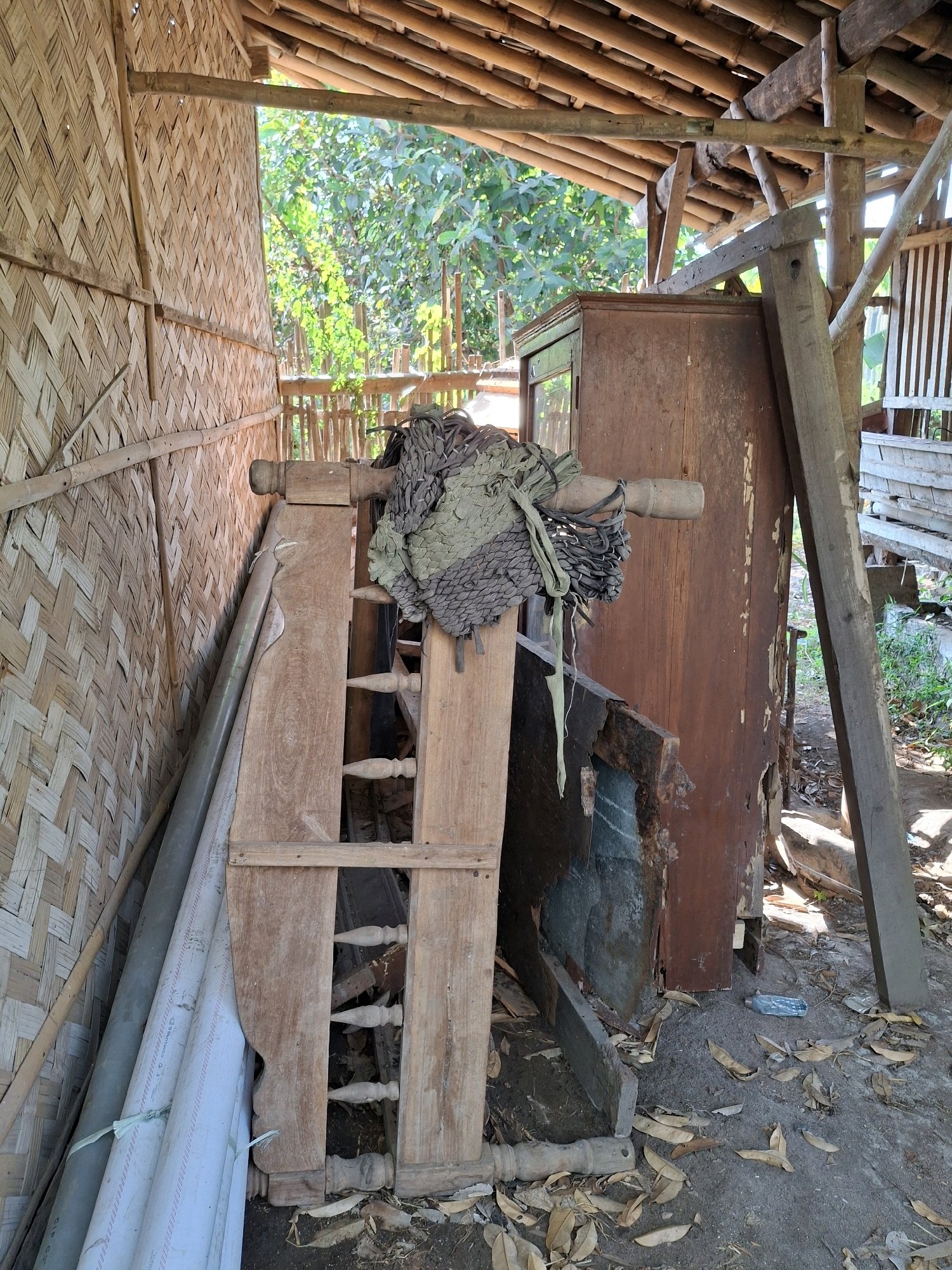 The image depicts an outdoor scene in Denok, Indonesia, likely a storage area or abandoned structure. The foreground features a jumble of objects, including several long PVC pipes, a damaged wooden cabinet with a glass door, and a piece of rustic wooden furniture. A piece of camouflage-patterned fabric is draped over the wooden furniture. The walls of the structure are made of woven bamboo, and the roof is constructed from bamboo poles. The ground is covered with dirt and fallen leaves. Sunlight filters through the open structure, illuminating the scene. In the background, trees and foliage are visible, suggesting a rural or semi-rural setting.