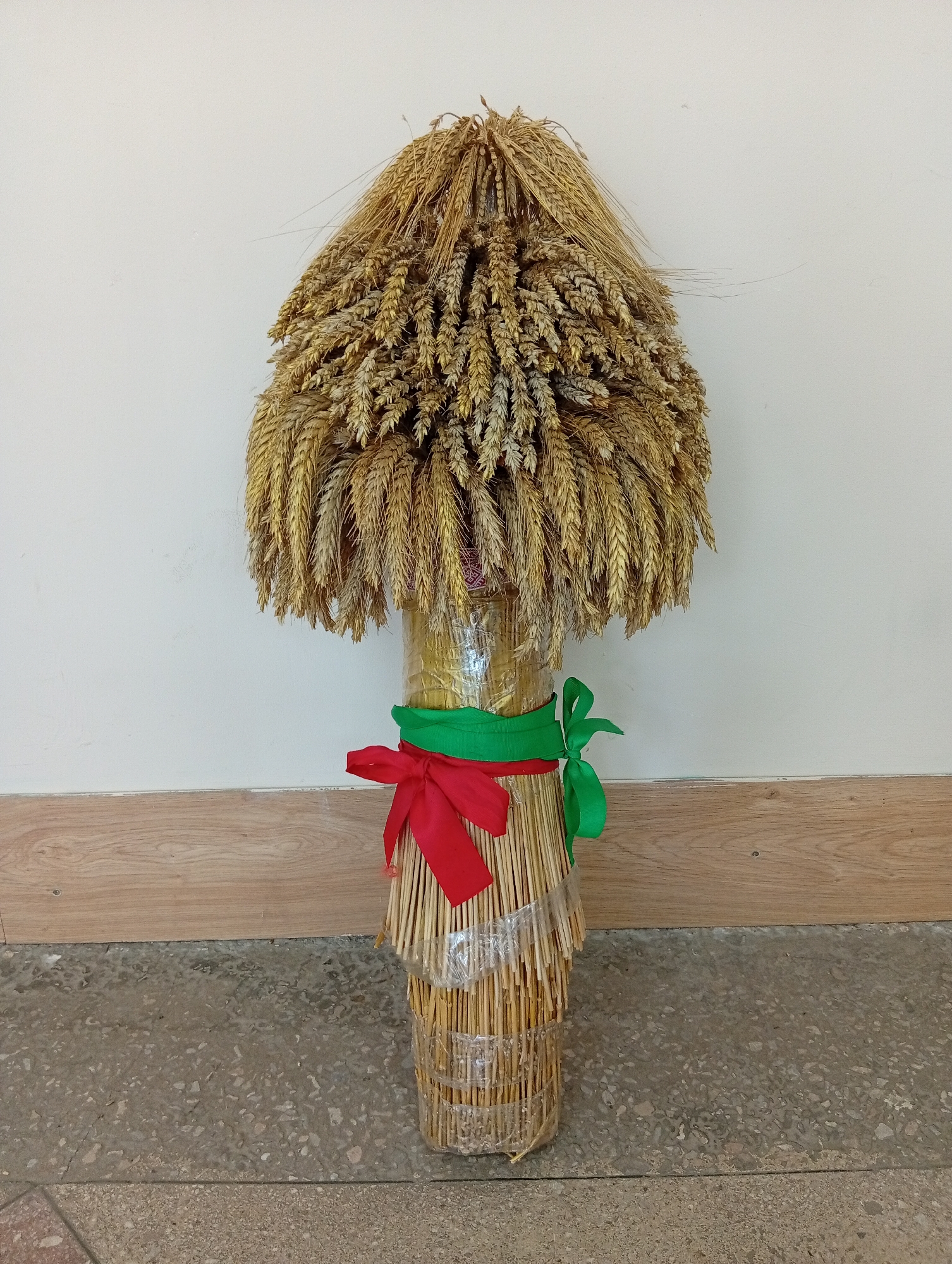 A decorative object made of wheat stalks and ears of wheat stands upright against a pale wall. The wheat is arranged in a conical shape at the top, with stalks hanging down to form a cylindrical base. A green ribbon is tied around the base, and a red ribbon is tied in a bow over the green ribbon. The base of the object is wrapped in clear plastic. The object is situated on a concrete floor with a wooden baseboard behind it. This scene is located in Крыўлянскі сельскі Савет, Belarus.