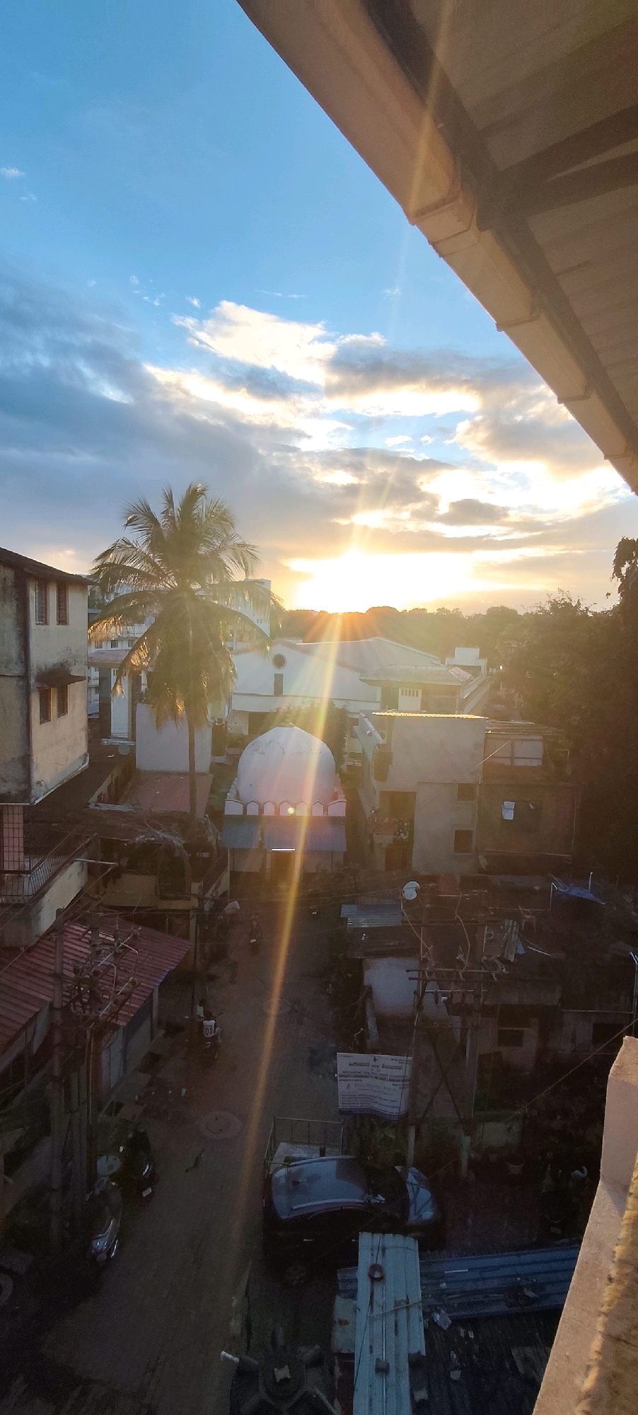 The image depicts a sunset scene in the city of Belagavi, India, viewed from a high vantage point. The sky is a mix of blue and orange hues, with clouds illuminated by the setting sun, creating visible sunbeams. In the foreground, a portion of a building's roofline is visible, casting a shadow over the scene.
Below the sky, a cluster of buildings forms the urban landscape. A prominent white dome structure, possibly part of a mosque or religious building, is centrally located. To its left, a tall palm tree stands out against the sky. The buildings are of various sizes and architectural styles, with some appearing to be older and others more modern. There are visible wires and electrical poles crisscrossing between the buildings, indicating an urban setting.
The lower portion of the image reveals a street with parked vehicles, including cars and motorcycles. Some structures appear to be shops or residences with makeshift roofing. The overall impression is a densely populated urban environment experiencing the golden hour of sunset.