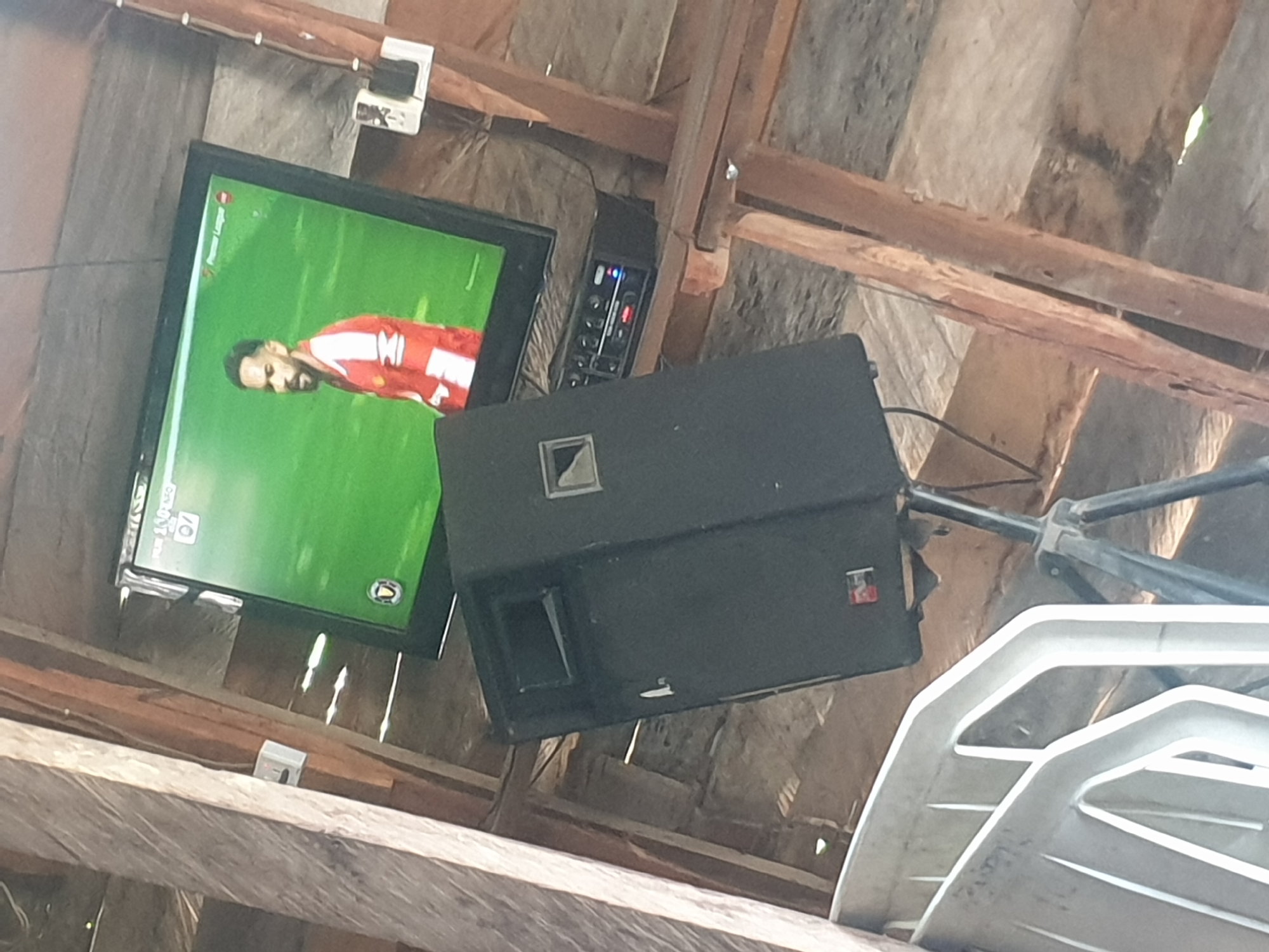 The image shows a TV playing a soccer game mounted on a wooden structure. The TV screen is green, and a player in a red and white jersey is visible. Next to the TV, there is an amplifier and a large black speaker on a stand. Below, there is a stack of white folding chairs. The location is unclear.