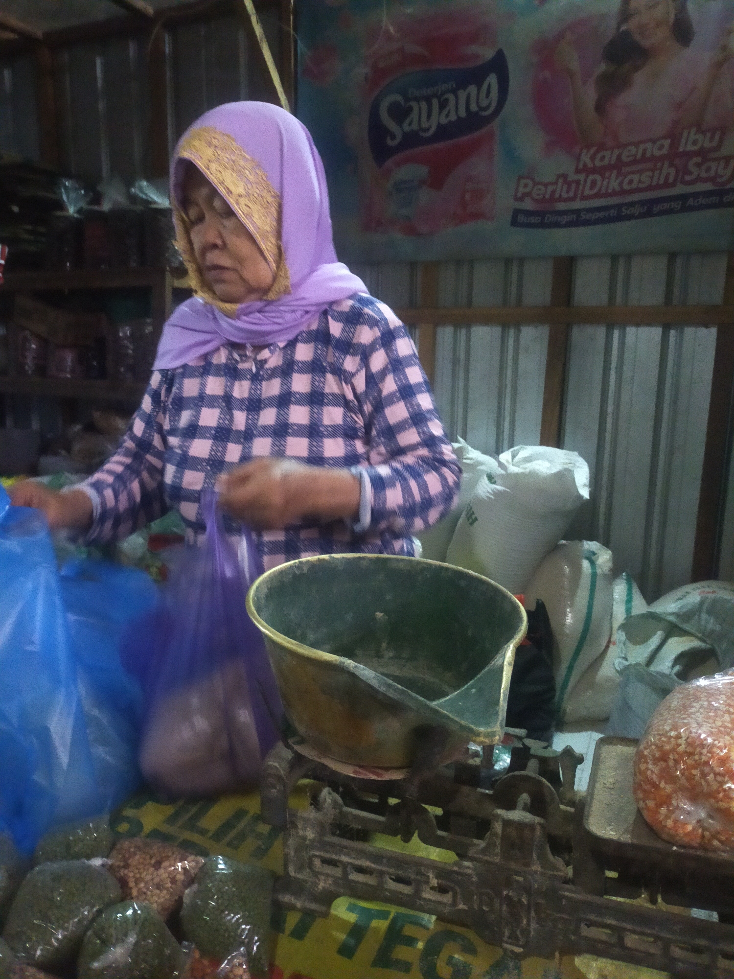 A middle-aged Indonesian woman, wearing a purple headscarf with gold trim and a blue and pink checkered blouse, stands in what appears to be a market stall in Trayeman, Indonesia. She is holding a purple plastic bag in her right hand and is looking down. In front of her is a vintage scale with a dark green basin on the left side. The scale rests on a yellow surface with green lettering, part of which is visible as "TEG...". Various bags of what appear to be food items, such as grains or beans, are visible around the scale. To the left of the woman are large blue plastic bags. The background consists of shelves with various items and a large advertisement poster featuring a woman and text in Indonesian. The lighting is dim and appears to be artificial.