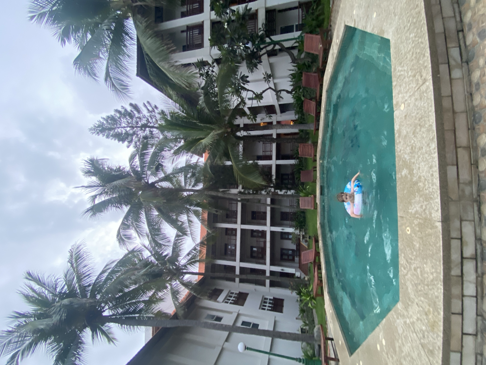 A sideways aerial shot shows a swimming pool next to a multistory white building surrounded by palm trees, in Phu Thuy Ward, Vietnam. A child floats in the pool in a multi-colored inflatable ring. Several empty wooden chairs and tables sit along the side of the pool closest to the building. The day appears cloudy.