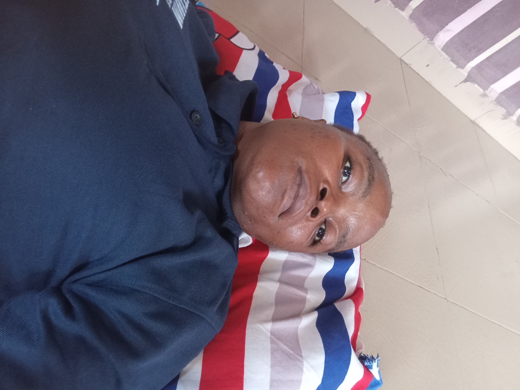 A person is lying on a bed with striped bedding, which is likely in a residential area of Kason, Nigeria. The person is wearing a dark blue collared shirt. The person is looking upwards, and their face is well-lit. The bedding is made up of horizontal stripes of red, white, and blue. The background shows a tiled floor.