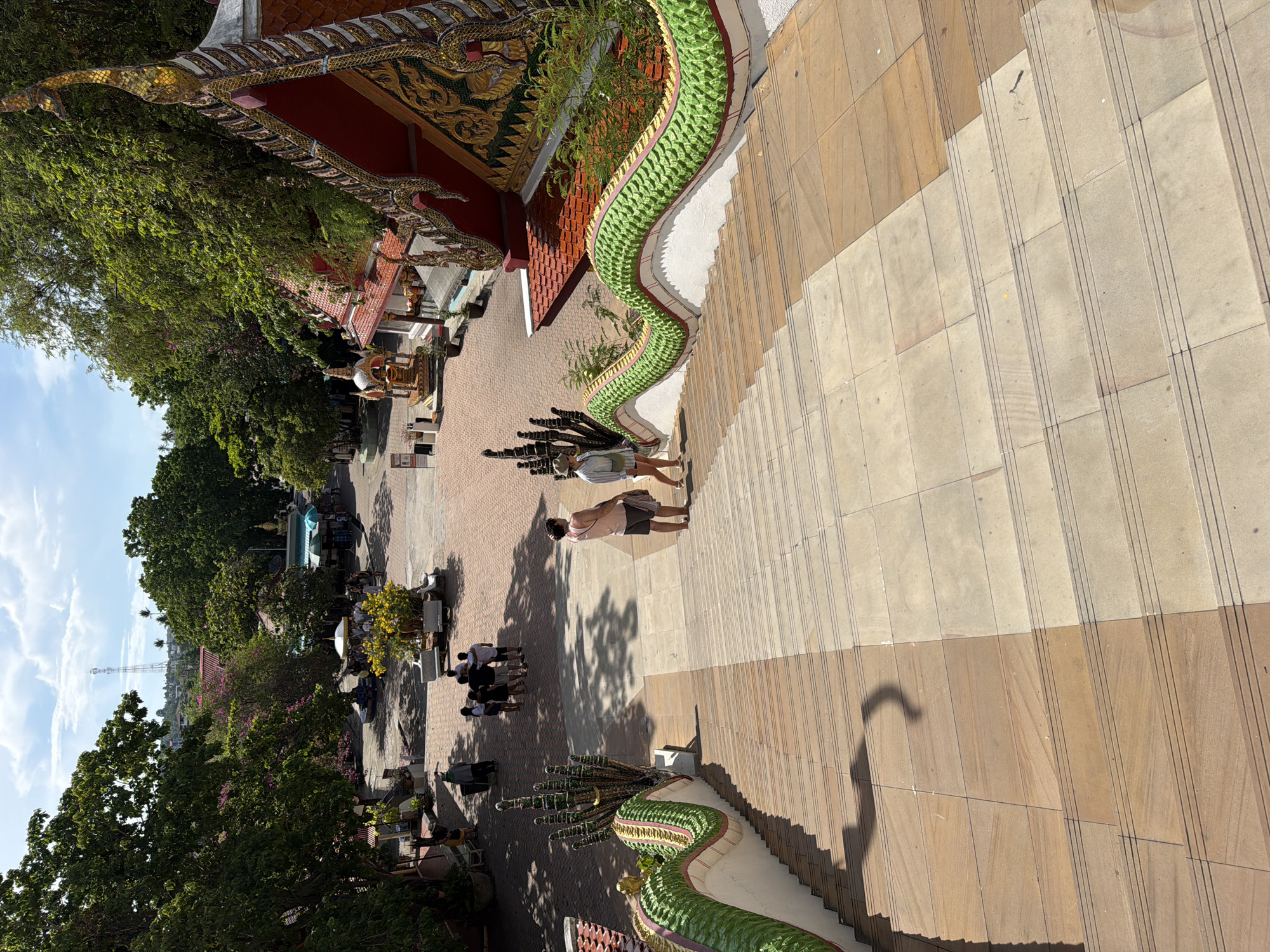 This image captures a view from the top of a wide, outdoor staircase adorned with ornate, green serpent railings, leading down to a temple courtyard in Baan Plai Laem, Thailand. The sun casts long shadows across the light-colored stone steps. On the staircase, two individuals are visible: a person wearing a pink shirt and dark shorts, and another person in a light-colored shirt and hat. They are facing each other, engaged in conversation or observation.
The courtyard below features a paved area with red brick patterns, shaded by lush green trees and vibrant pink and yellow flowering plants. Several people are gathered in small groups, some standing and others sitting, in the open courtyard. A white umbrella provides shade for a group of people.
To the right of the staircase, a section of a temple building with a red roof and intricate golden detailing is visible, its architectural style characteristic of Thai temples. In the background, the cityscape of Baan Plai Laem unfolds with buildings and a prominent communication tower piercing the bright blue sky, dotted with white clouds. The overall scene depicts a sunny day at a cultural landmark, with visitors and locals interacting within the temple grounds.