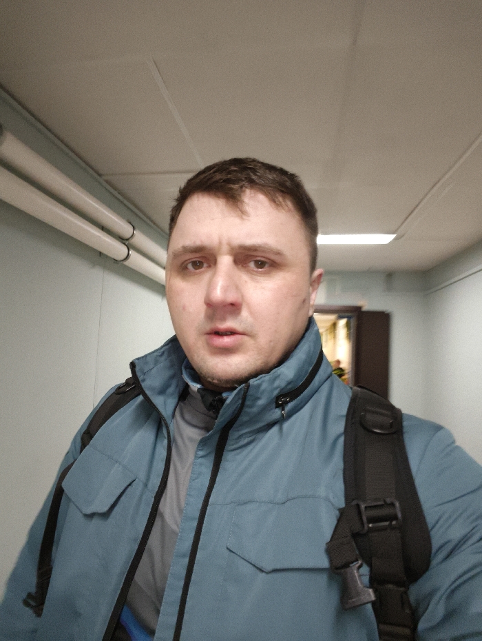 A male individual is depicted in a chest-up frontal shot, likely a selfie, within an indoor hallway in the city of Im. Gastello, Russia. He has short, dark brown hair and light eyes, with a neutral expression and slightly parted lips. He wears a teal or blue-green jacket with black zippers and shoulder accents, over a grey long-sleeved shirt. A black backpack strap is visible over his right shoulder. The background features a light-colored wall on the left, with two parallel fluorescent tube light fixtures mounted horizontally. The ceiling is white or off-white with visible seams. Further down the hallway, a rectangular overhead light fixture is visible, illuminating the space. On the far right, a partially visible doorway reveals an interior space with warm lighting and a glimpse of what appears to be wooden elements. No other individuals are present.