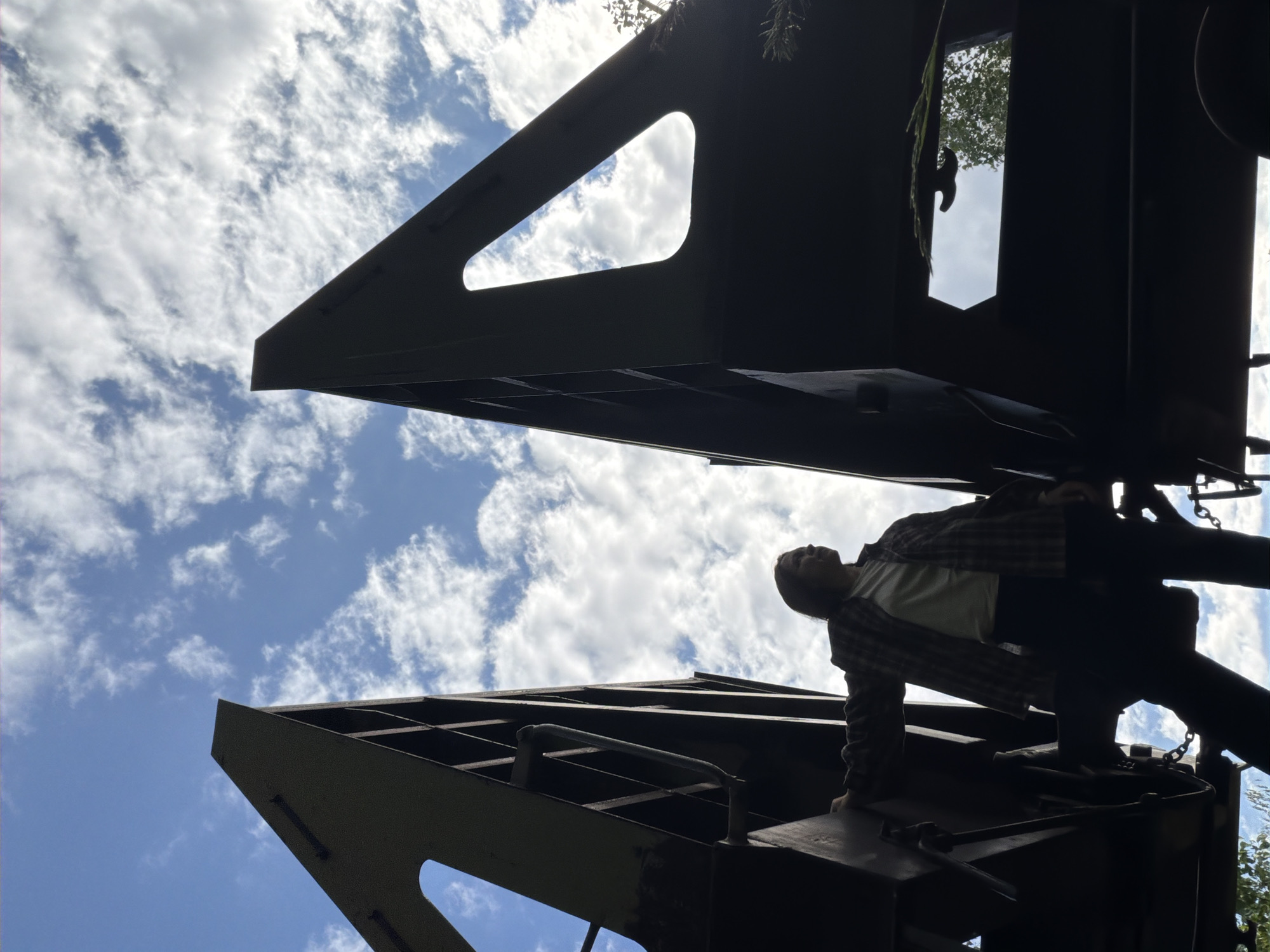 This low-angle photograph captures a person on a large, dark, angular metal structure against a bright blue sky filled with scattered white clouds. The structure, possibly a bridge or industrial machinery, is seen from below, forming a stark silhouette that dominates the foreground and midground. Its design features geometric cut-outs and internal bracing.
On the right side of the frame, a person, mostly in silhouette, is visible. They appear to be standing on or climbing the metal edifice, leaning forward with their head tilted slightly upwards. They are wearing a plaid shirt over a lighter undershirt, dark pants, and shoes. Chains are visible near their feet on the structure. Some green foliage from trees can be seen at the top right, peeking over the metalwork.
The scene takes place outdoors during the daytime, under bright, cloudy conditions. The dramatic perspective emphasizes the scale and imposing nature of the metal structure. The location context is Chaikovsky, Russia. No visible text is present in the image.
