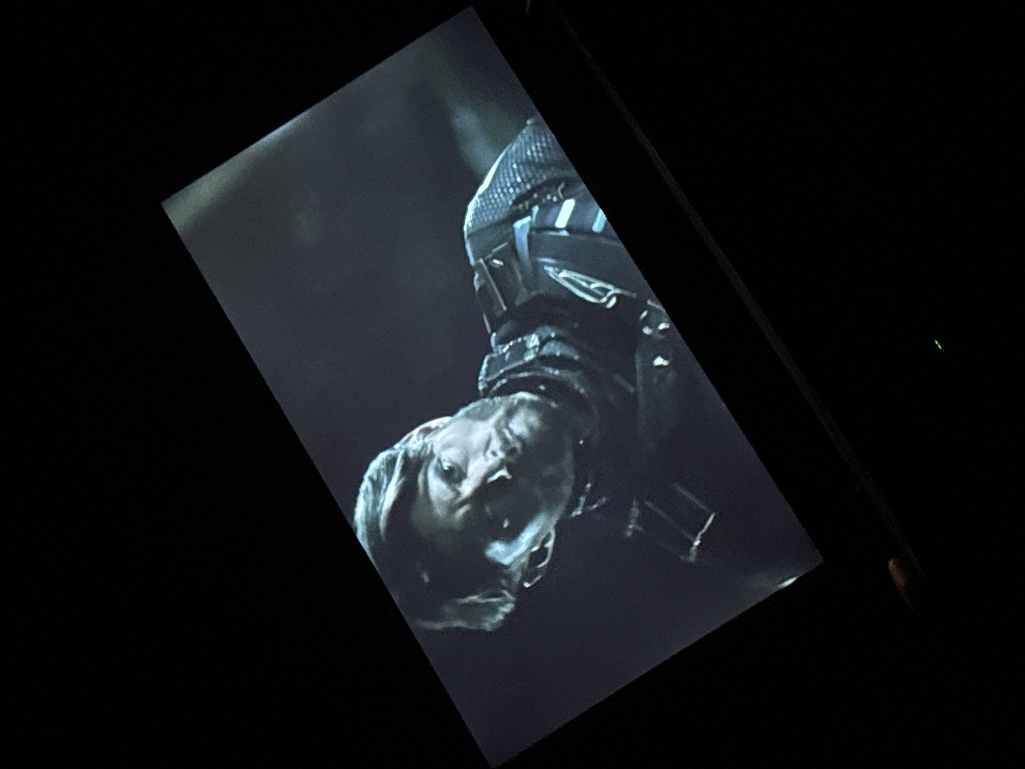 A man with a beard and short, dark hair is shown from the chest up, looking slightly down and to his right. He is wearing dark clothing with a textured, possibly armored, upper portion. The image is a photograph of a screen, which is angled and appears to be dark in its surroundings. There is no visible indication of the city of Dnipro, Ukraine.