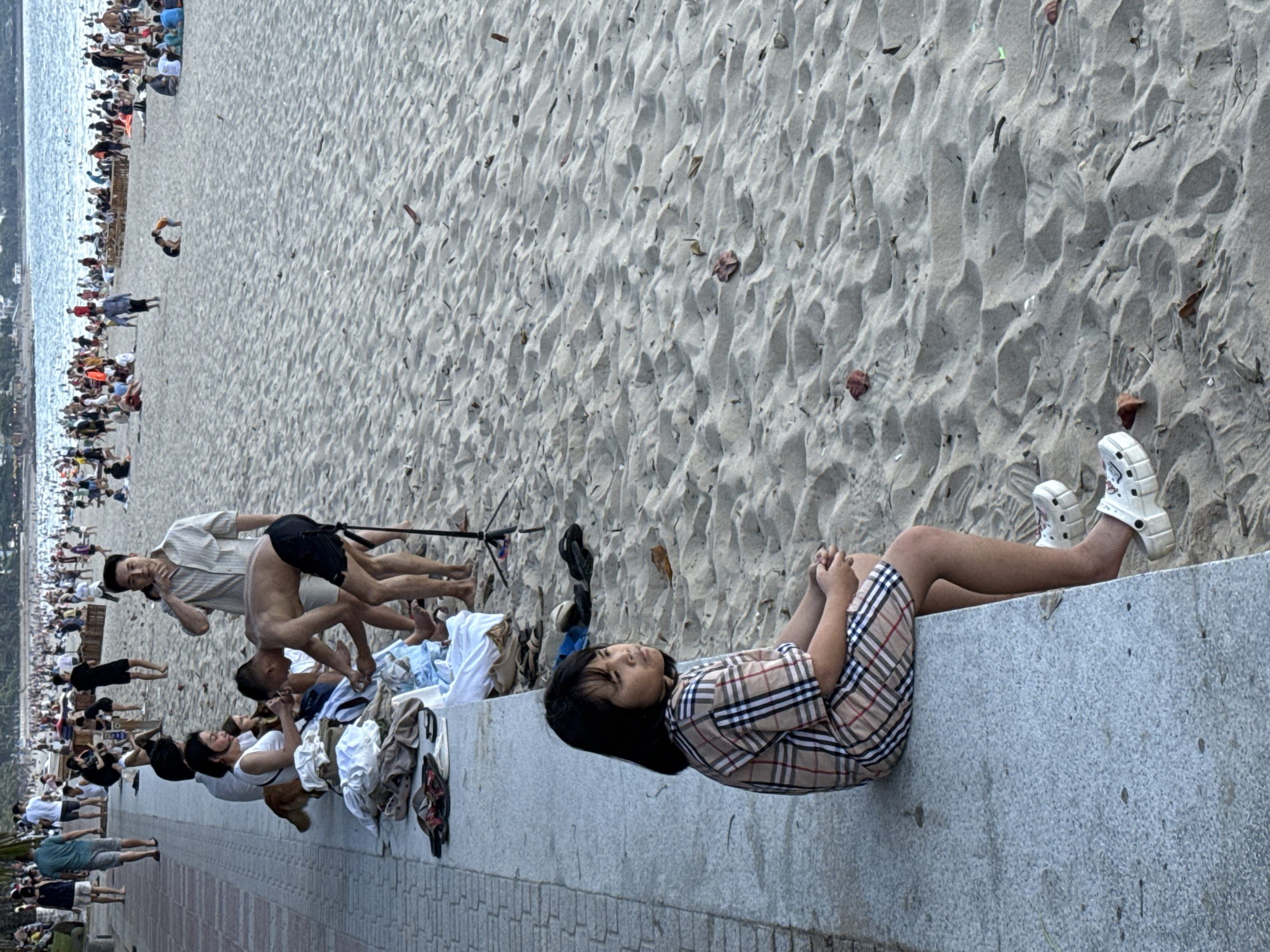 A young Vietnamese girl with dark hair is seated on a concrete wall overlooking a sandy beach in Nha Trang Ward, Vietnam. She is wearing a tan and plaid patterned dress and white platform Crocs. Her hands are clasped in her lap, and she is looking to her right. Behind her, several people are engaged in various activities on the beach. In the middle ground, a man in a light-colored, short-sleeved shirt and dark shorts is bent over, appearing to interact with someone or something on the sand. A woman in a white tank top is seated next to him on the wall, also looking towards the beach. A brown dog is visible near them. Further back, many people are scattered across the beach, some sitting, some standing, and some walking near the water. The beach is covered in fine, light-colored sand, with some debris and footprints visible. The ocean is in the background, with gentle waves. The overall scene depicts a lively beach atmosphere with people enjoying a day by the sea.