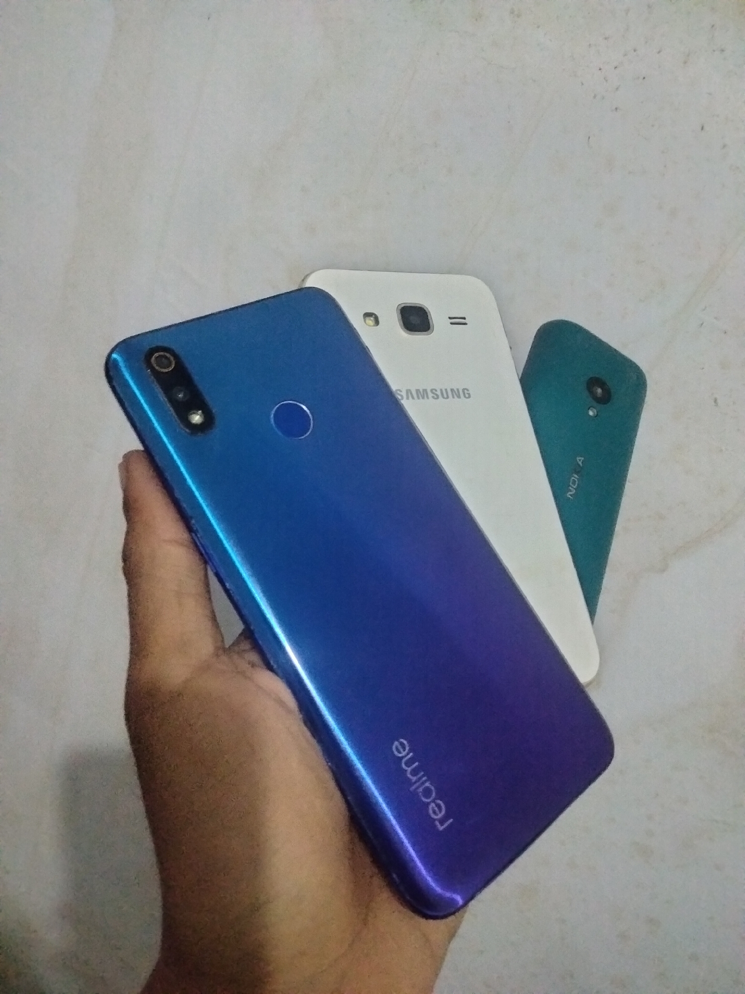 A hand holds a blue smartphone with a gradient finish, displaying the "realme" logo. Behind it, a white Samsung smartphone is visible, with the "SAMSUNG" logo printed on its back. Partially obscured by the Samsung phone is a teal Nokia phone, with the "NOKIA" logo visible. The phones are arranged on a textured, light-colored surface, likely a table or floor, with the overall scene appearing to be indoors. There is no information in the image to suggest a specific location in Antirogo, Indonesia.
