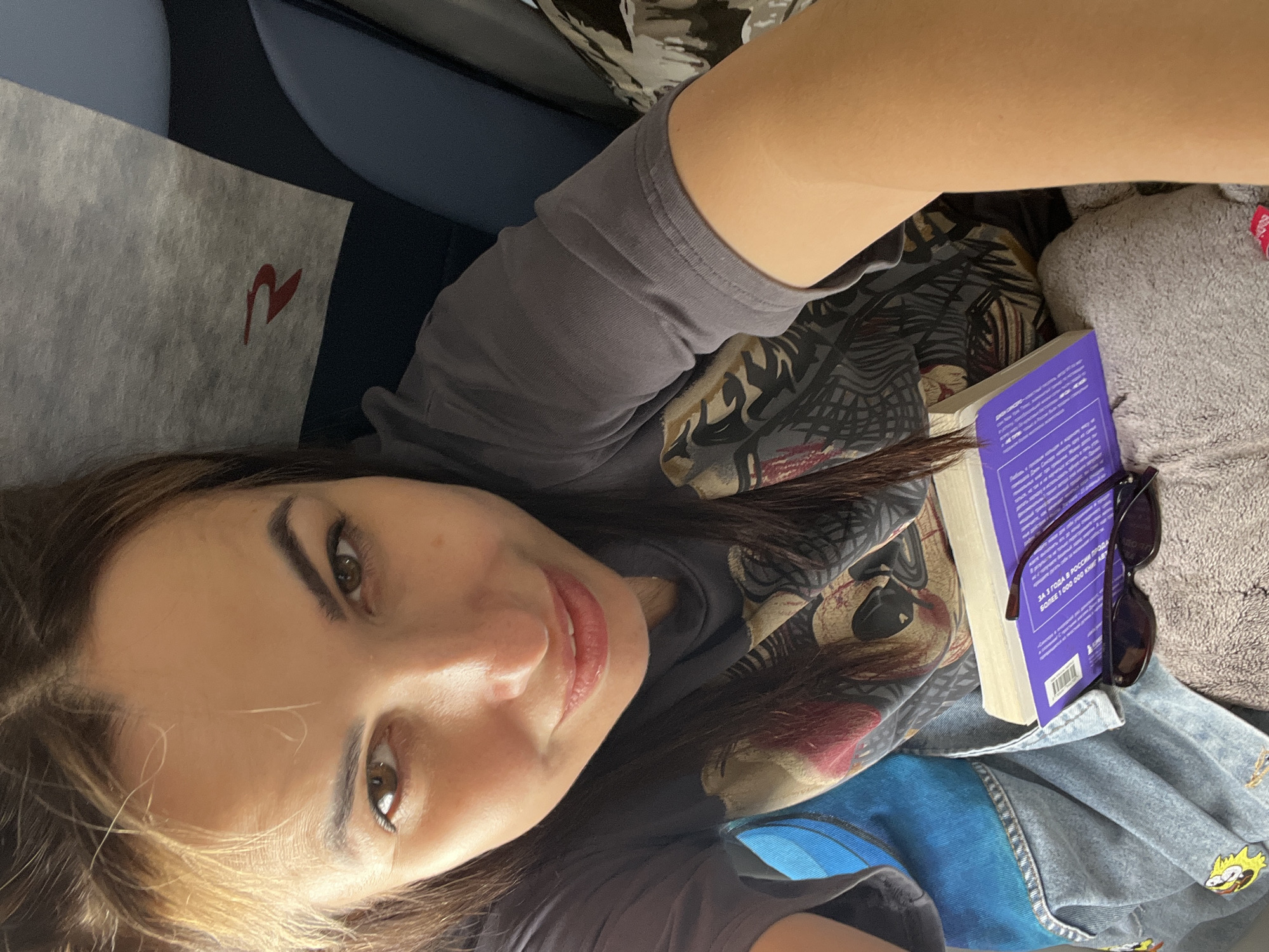 A woman is lying down on a surface, possibly a seat, with her head tilted up and looking at the camera. She has long, brown hair, brown eyes, and is wearing a dark grey t-shirt with a graphic print. Her left arm is visible and appears to be resting on her chest. In front of her, resting on a grey, textured surface, is a book with a purple cover and a pair of dark sunglasses placed on top of it. The background suggests an indoor setting, possibly a mode of transportation, with a hint of blue seating and a grey fabric with a red logo visible behind her head.