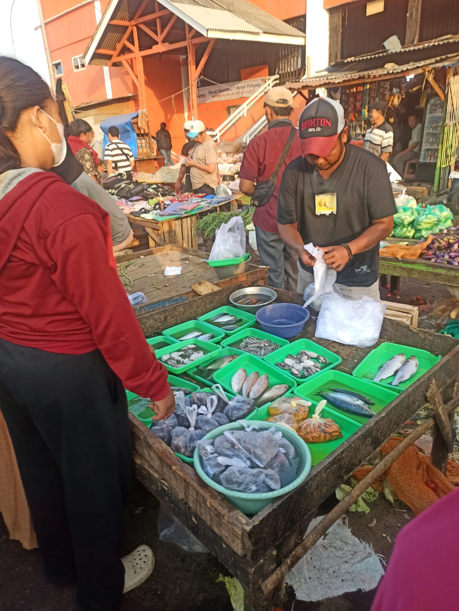 A busy street market in Cikarang, Indonesia, is captured from a slightly elevated, eye-level perspective. In the foreground, a person wearing a red hoodie and black pants stands with their back to the camera, facing a wooden stall laden with various goods. A man in a dark t-shirt and a baseball cap with "BRIXTON MFG. CO." embroidered on it, stands behind the stall, actively engaged in packaging a white plastic bag.
The stall itself is a vibrant display of fresh produce and seafood. Numerous green plastic trays are filled with fish of various sizes and types, some whole and some in plastic bags. There are also containers with what appear to be shellfish. Behind the fish stall, other vendors are visible, with stalls selling a variety of items, including vegetables like chilies and eggplant, as well as other packaged goods.
The environment suggests a bustling marketplace. In the background, buildings with orange facades and tiled roofs indicate urban infrastructure. Other people are present, some browsing, others working, adding to the dynamic atmosphere of the scene. The overall lighting suggests it is daytime, likely morning or late afternoon, given the warm, natural light. The ground is unpaved and appears to be dirt or gravel, typical of many outdoor markets.