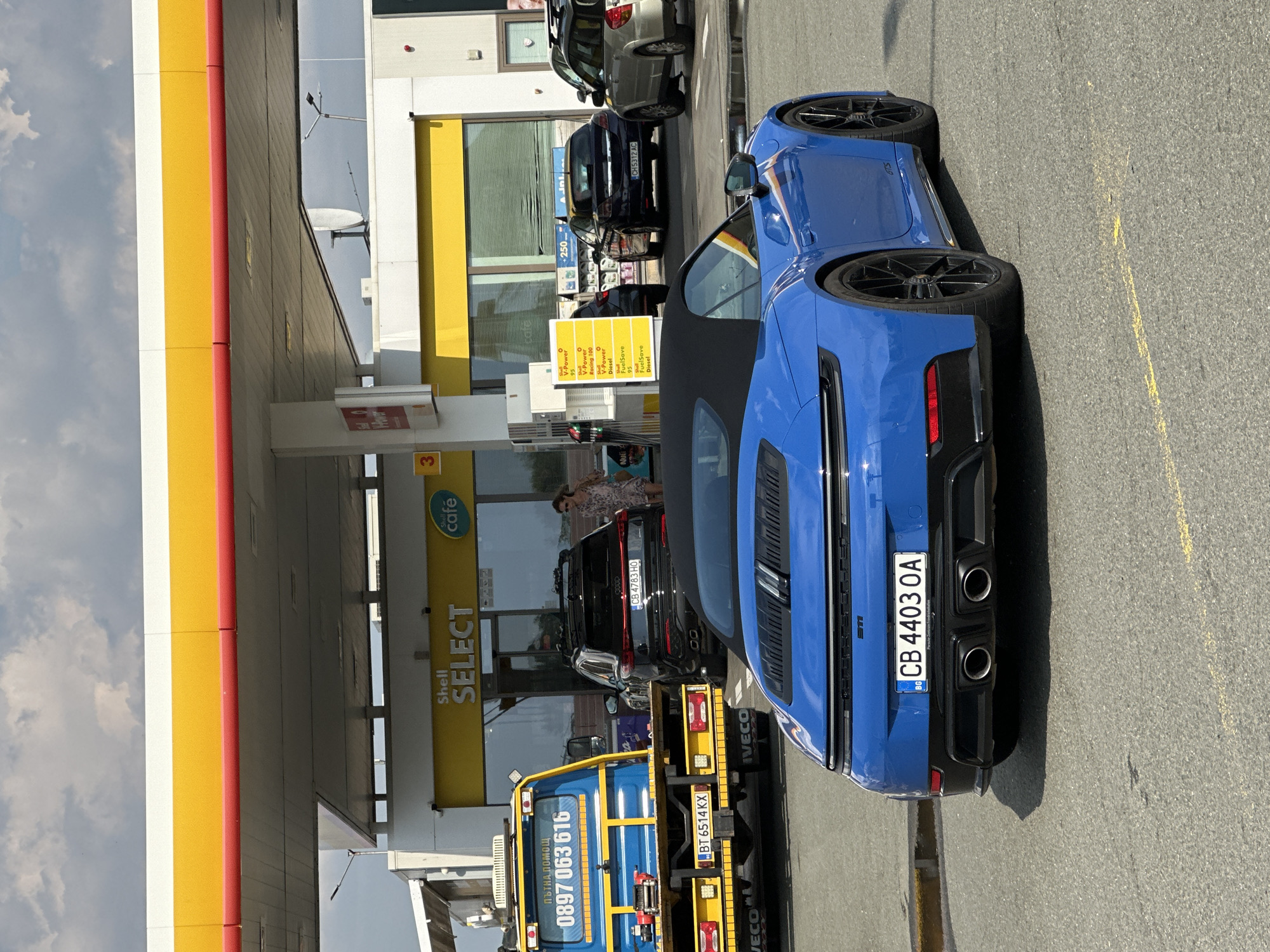 The image depicts a blue Porsche 911 convertible parked at a Shell gas station in Drazhevo, Bulgaria. The car's license plate reads "CB 4403 OA". In the background, a blue tow truck with the phone number "0897 063 616" is visible, along with a black SUV and a silver station wagon. A woman in a patterned dress is walking near the gas station entrance. The gas station features Shell Select and Shell Cafe branding. The sky is overcast with clouds.
