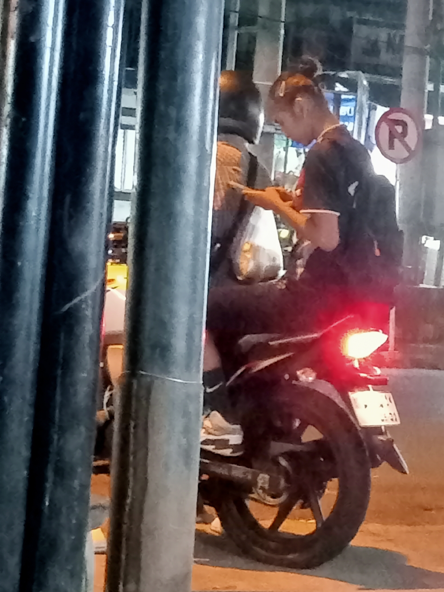 The image captures a nighttime scene on a street in Mangunsari, Indonesia. Two individuals are seated on a motorcycle. The person in the foreground, a woman with her hair pulled back in a bun, is looking down at her mobile phone and appears to be holding it with both hands. She is wearing a dark t-shirt and a backpack. Behind her, another person is partially visible, wearing a helmet. The motorcycle's brake light is illuminated red. A "No Parking" sign with a red circle and diagonal line over the letter "R" is visible in the background, indicating a traffic regulation. The environment appears to be an urban setting with some visible streetlights and possibly buildings in the distance, though they are out of focus. The ground is paved and illuminated by ambient light.